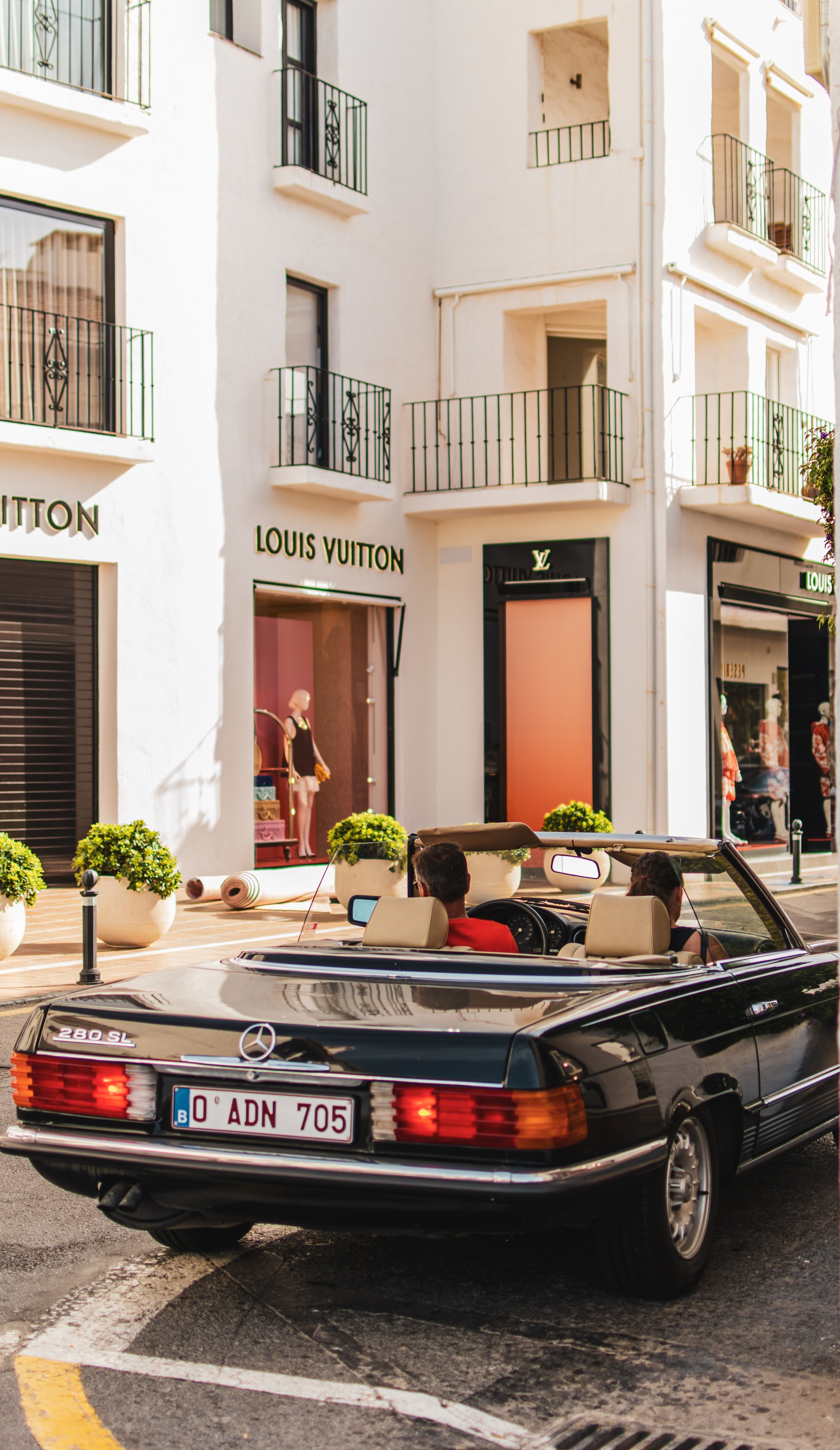 Vintage Mercedes convertible in front of a Louis Vuitton store with mannequins in the window display, on a sunny street.