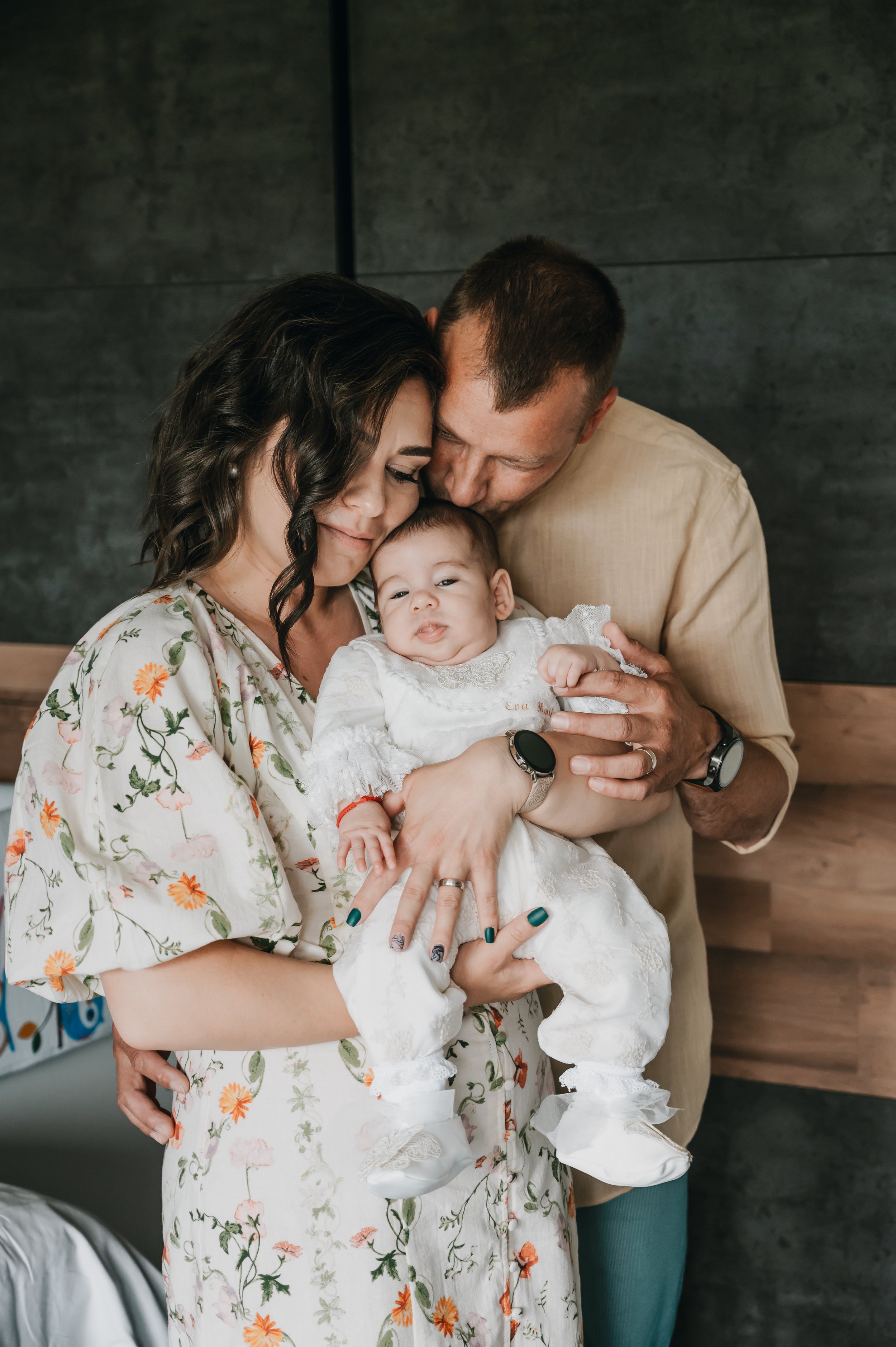 A couple lovingly holding their baby in a floral dress, with the father kissing the baby's head. The family is close and affectionate, capturing a tender moment.