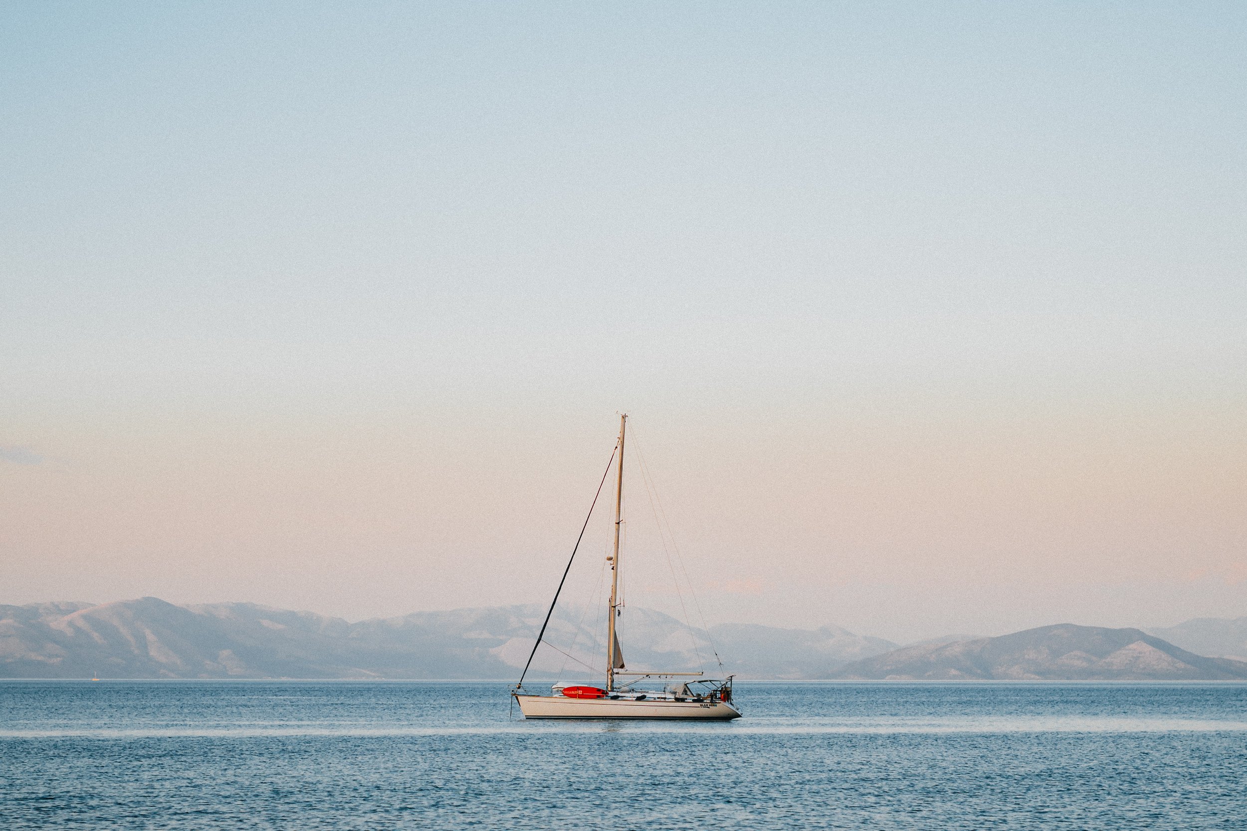 Sailboat on calm water with distant mountains under a clear sky.