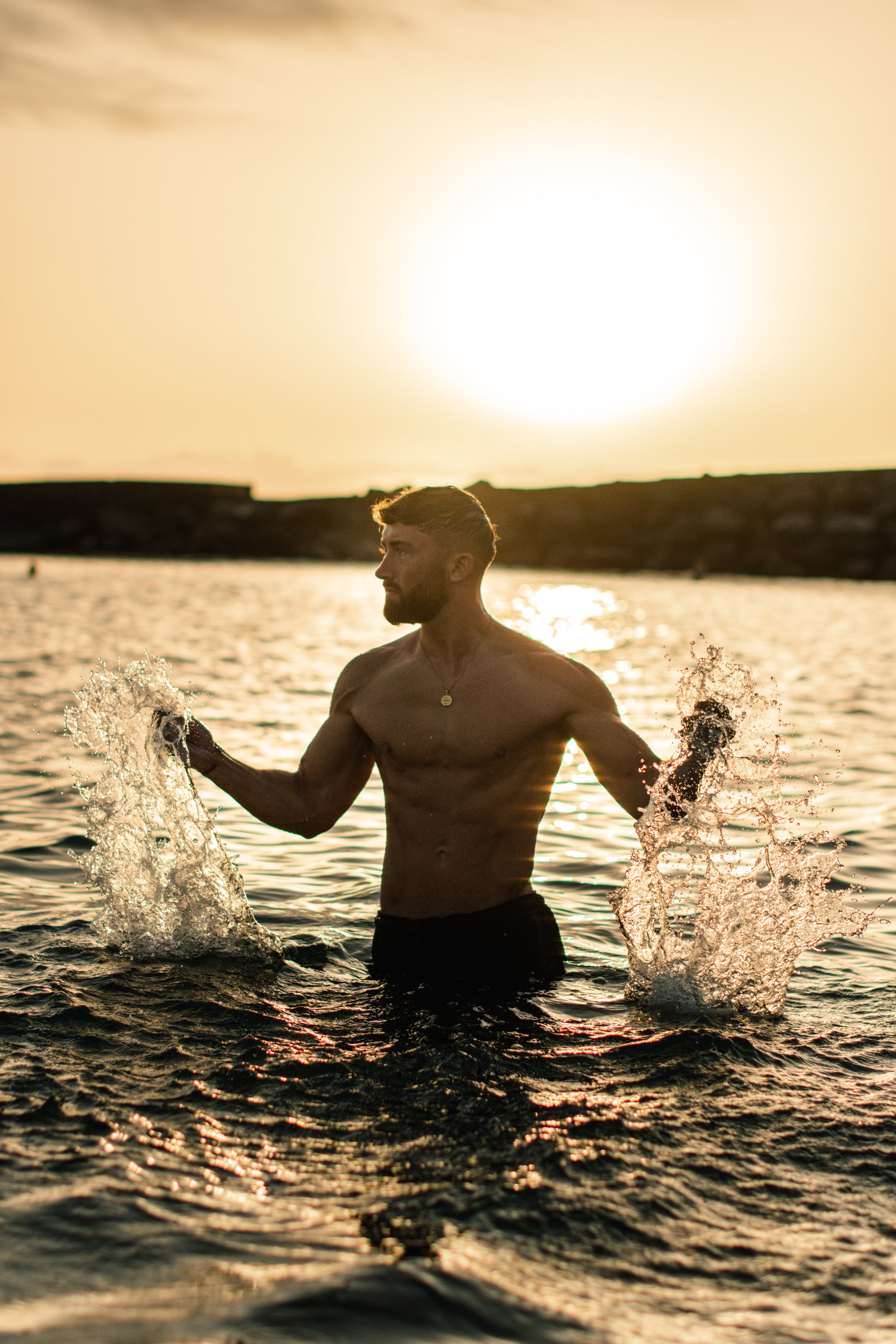 A shirtless man with a necklace stands in the water at sunset, splashing waves around him.