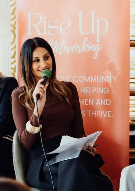 A woman in a black dress standing in front of a red banner that reads 'Rise Up Networking' and mentions supporting ambitious women and fundraising for Yorkshire Children's Charity.