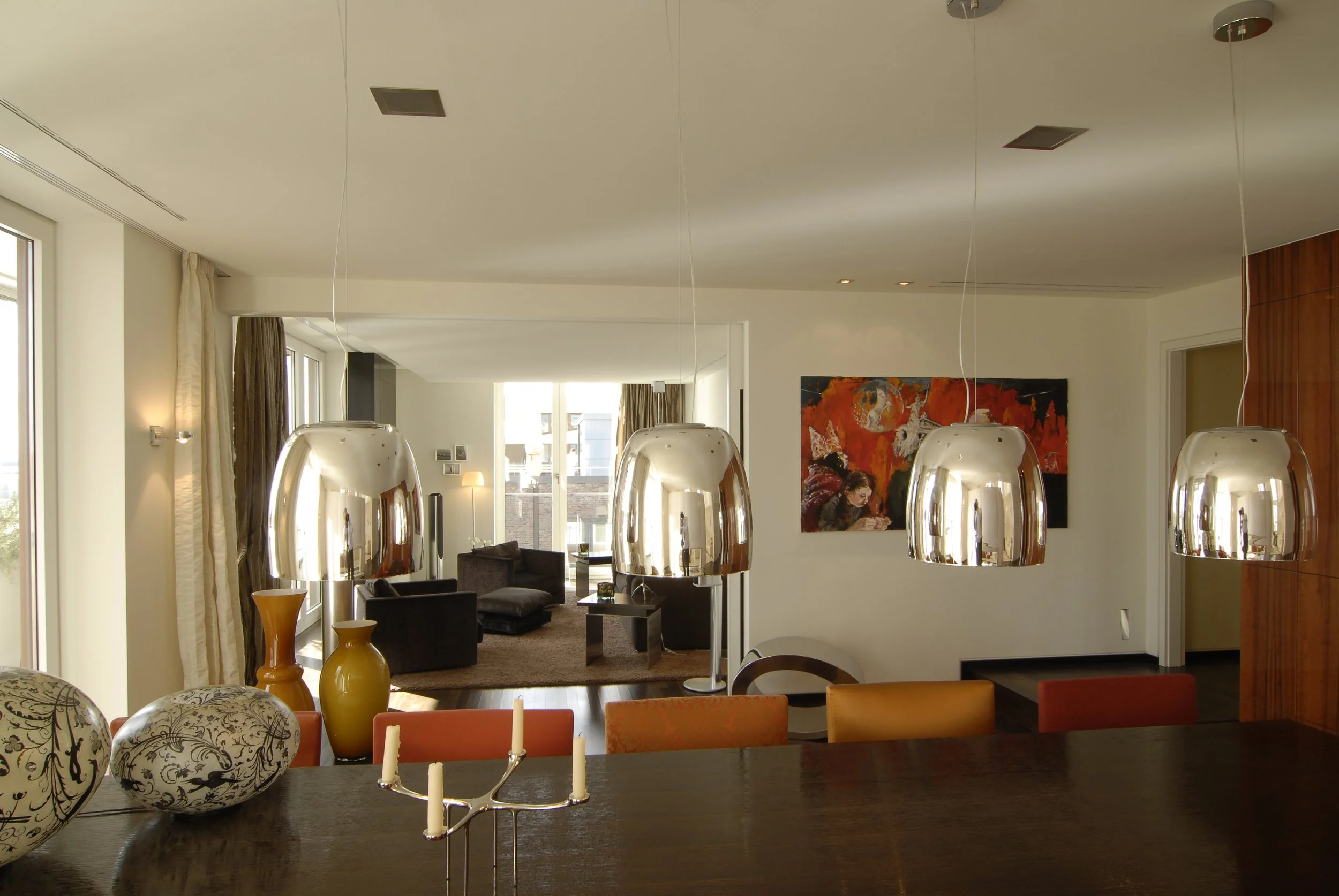 Moderner Essbereich mit vier hängenden silbernen Lampen, einem dunklen Holztisch, Kerzenhalter, Vasen und keramischen Dekorationen. Im Hintergrund ein Wohnzimmer mit Sitzmöbeln, einem Teppich und einem bunten Wandbild.