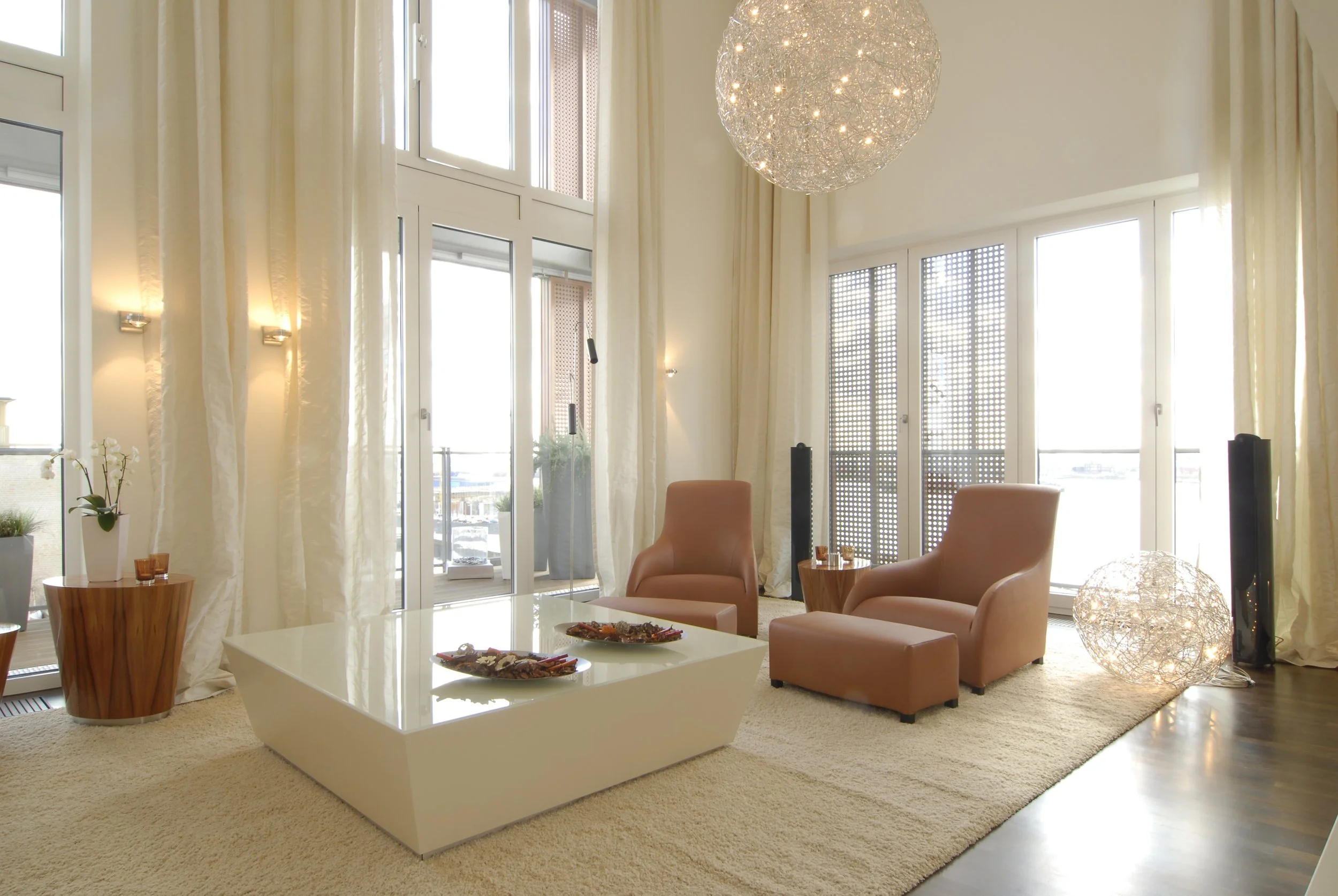 Ein helles Wohnzimmer mit großen Fenstern, eleganten beige Sessel, einem weißen Couchtisch mit Dekoration, einem großen beleuchteten Kronleuchter und Sonnenschutzjalousien.