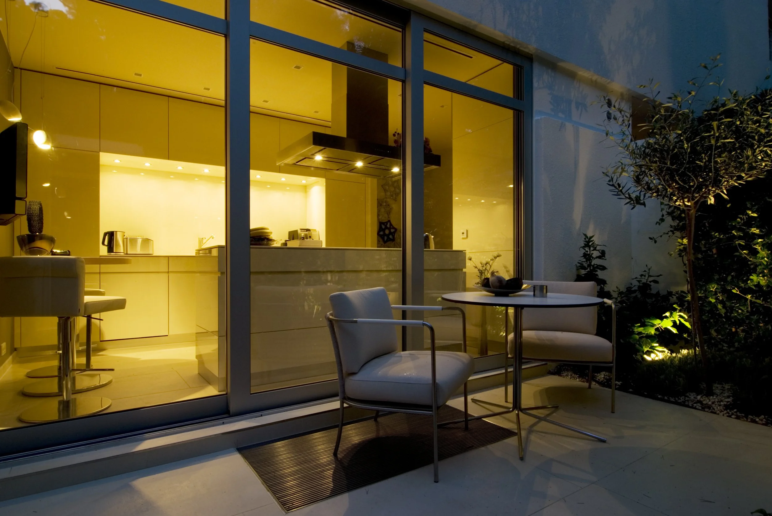 Blick auf die Terrasse eines modernen Hauses bei Nacht, mit einem kleinen Tisch und zwei Stühlen im Außenbereich, Blick durch eine große Glasfassade in die gut beleuchtete Küche.