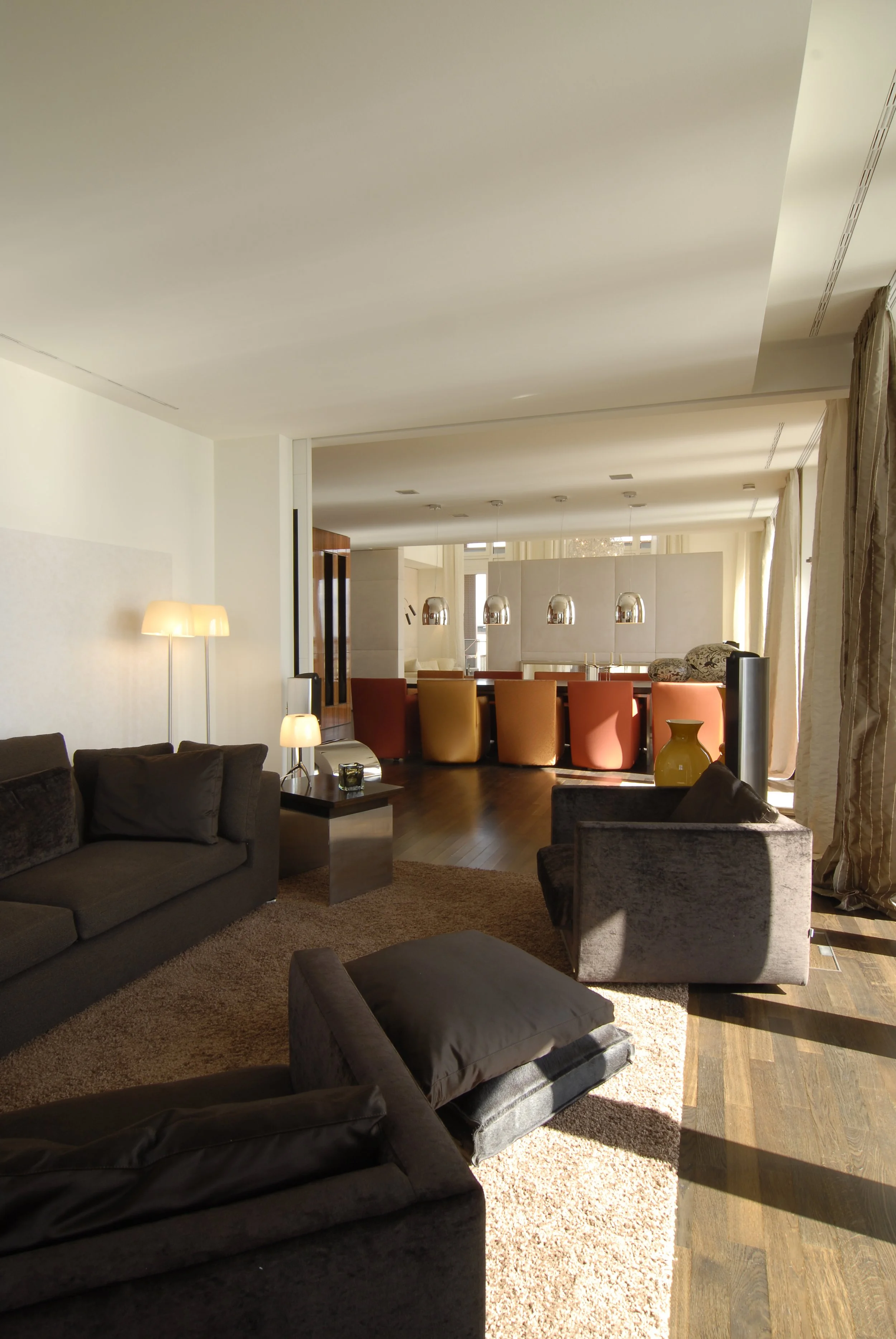 Moderner Wohnzimmerbereich mit dunklen Sofas, einem kleinen Beistelltisch, Stehlampen, einem Teppich, einem Drehstuhl, einem Vasen und einem Essbereich im Hintergrund mit Stühlen und Pendelleuchten.
