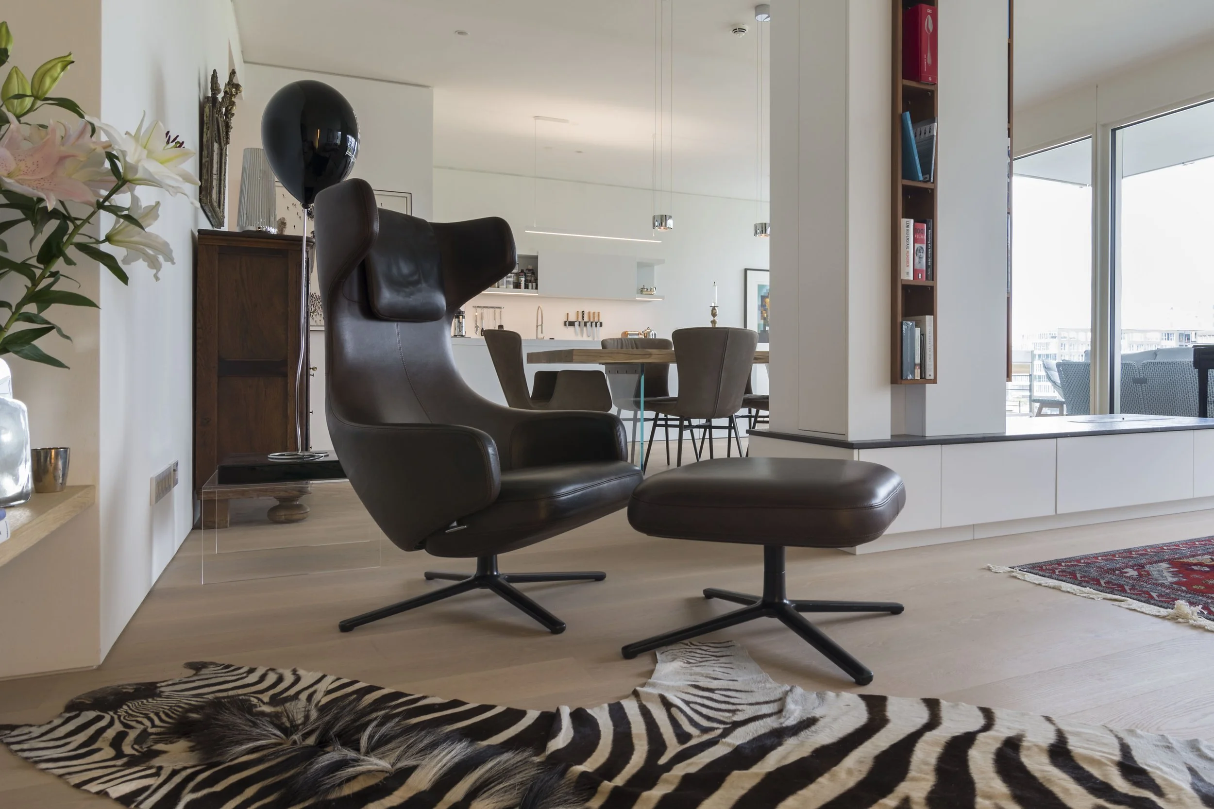 Modernes Wohnzimmer mit schwarzem Designer-Sessel und Hocker, Zebra-Fell-Teppich, offene Küche im Hintergrund, große Fenster mit Balkonblick.