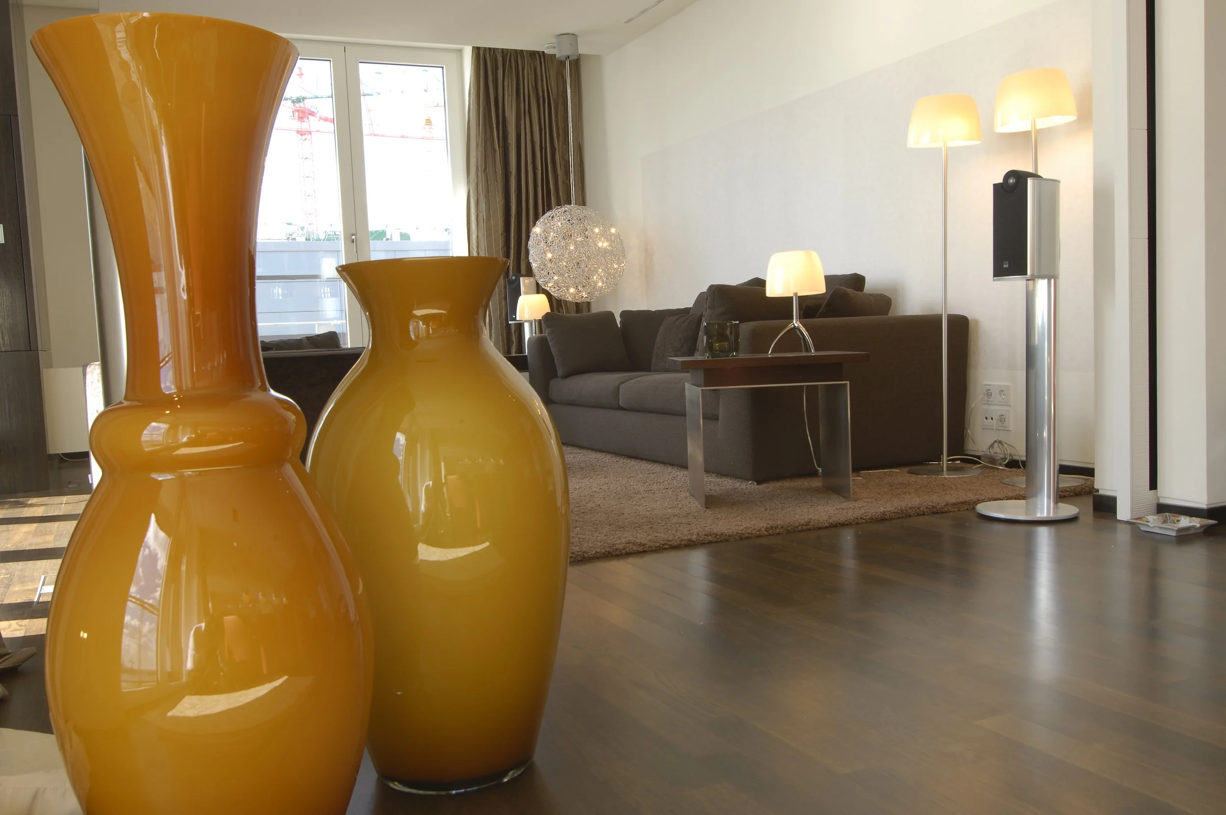 Wohnzimmer mit dunklem Sofa, Stehlampen, einem modernen Heizstrahler, einem Teppich und dekorativen Vasen im Vordergrund, große Fenster mit Vorhängen.