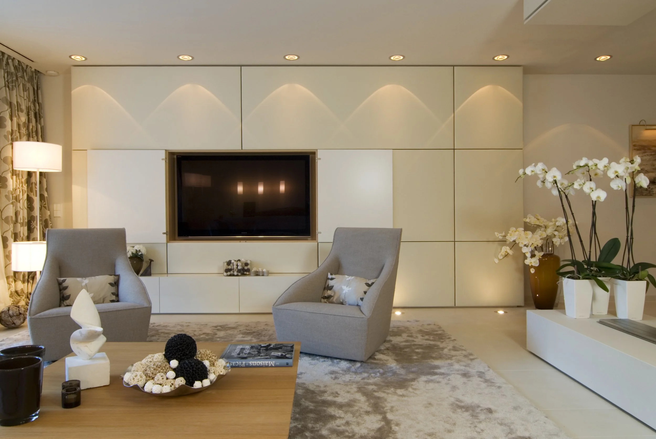 Moderner Wohnzimmerbereich mit weißen Wandschrank, Fernseher, zwei grauen Sessel, Holztisch mit Dekorationen, und weißen Orchideen in großen Vasen.