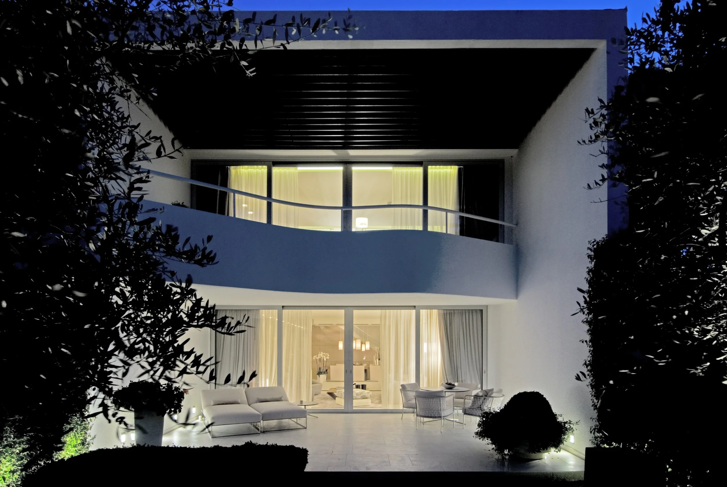 Modernes, beleuchtetes Haus mit Balkon in der Nacht, weiße Wände, große Fenster, Gartenmöbel im Außenbereich, Bäume und Büsche