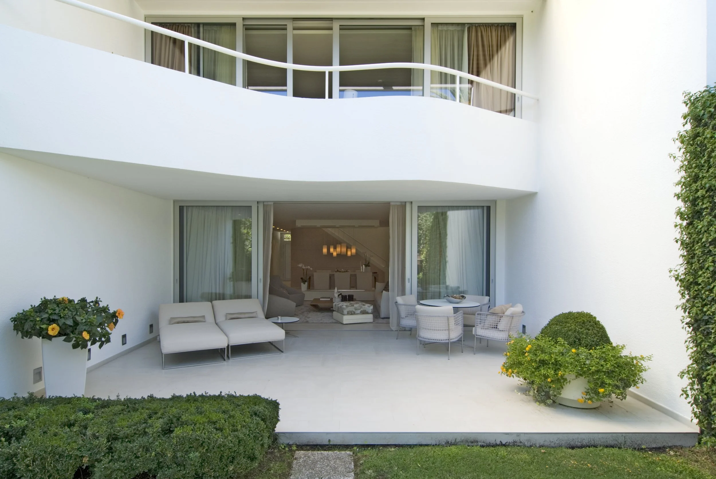 Ein moderner Balkon mit weißen Möbeln, einem runden Tisch, zwei Liegen, bunten Pflanzen und einem Blick ins Wohnzimmer dahinter.