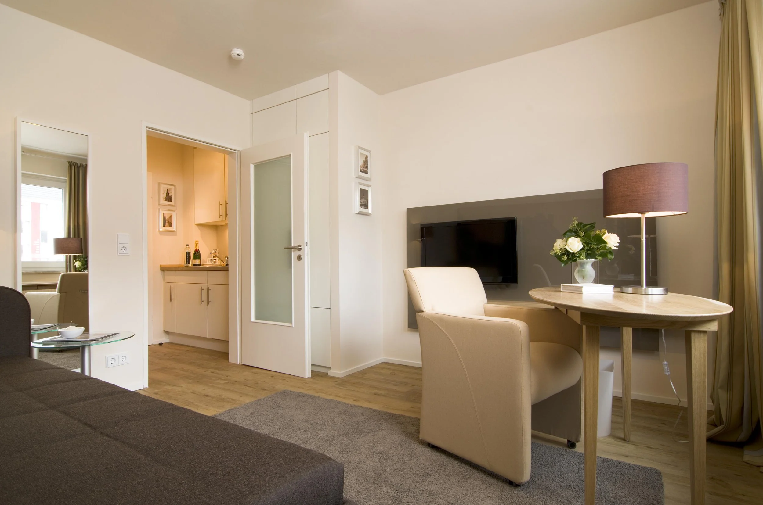 Innenaufnahme eines modernen Hotelzimmers mit Sitzbereich, Fernseher, Tischlampe, Blume und dunkelbraunem Sofa, mit Blick auf eine offene Küche und Fenster.