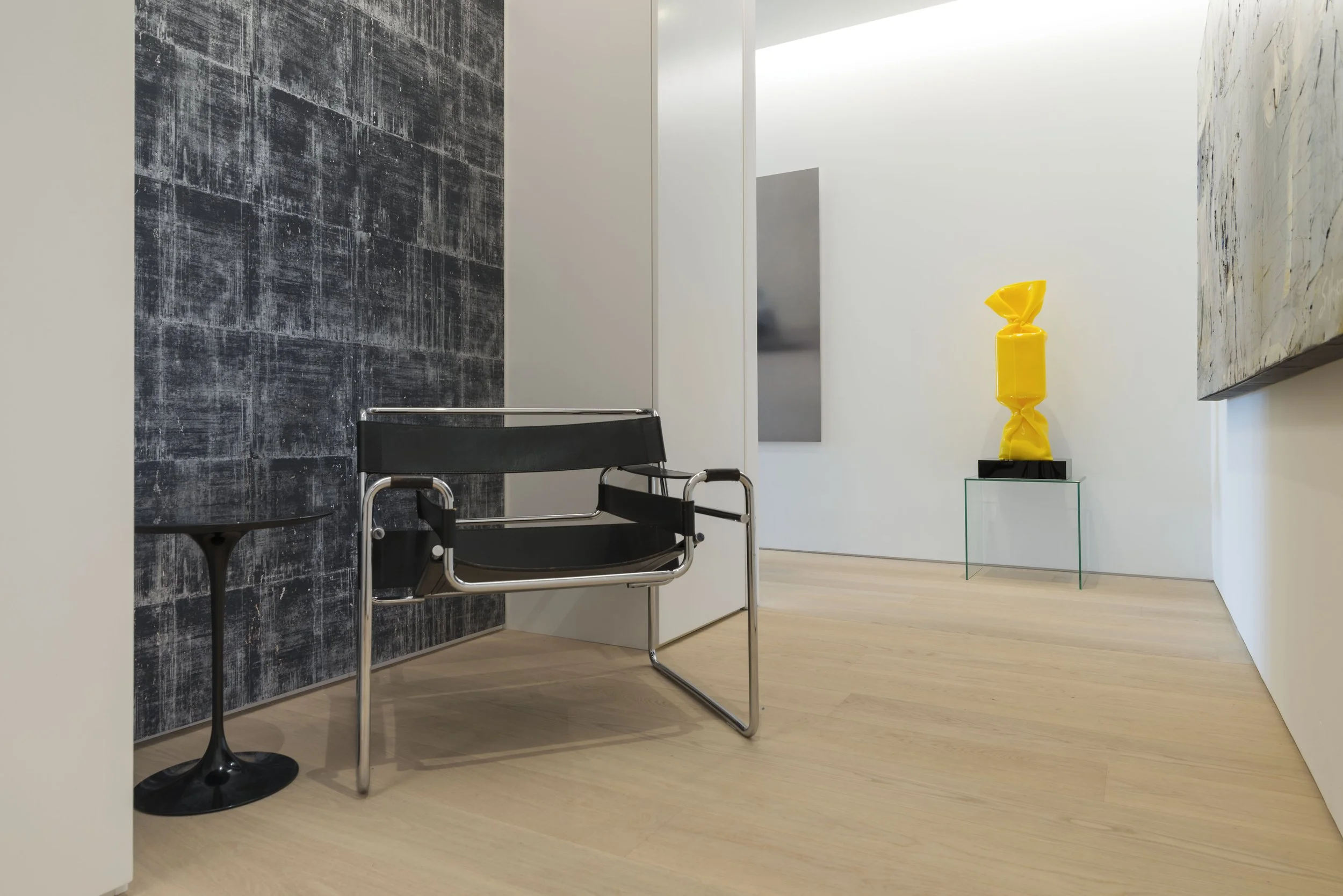 Innenraum einer modernen Kunstgalerie mit schwarzem Sessel, schwarzem Beistelltisch, grauer Wand hinter dem Sessel, weißen Wänden und einem Glasständer mit gelber Skulptur auf der rechten Seite.