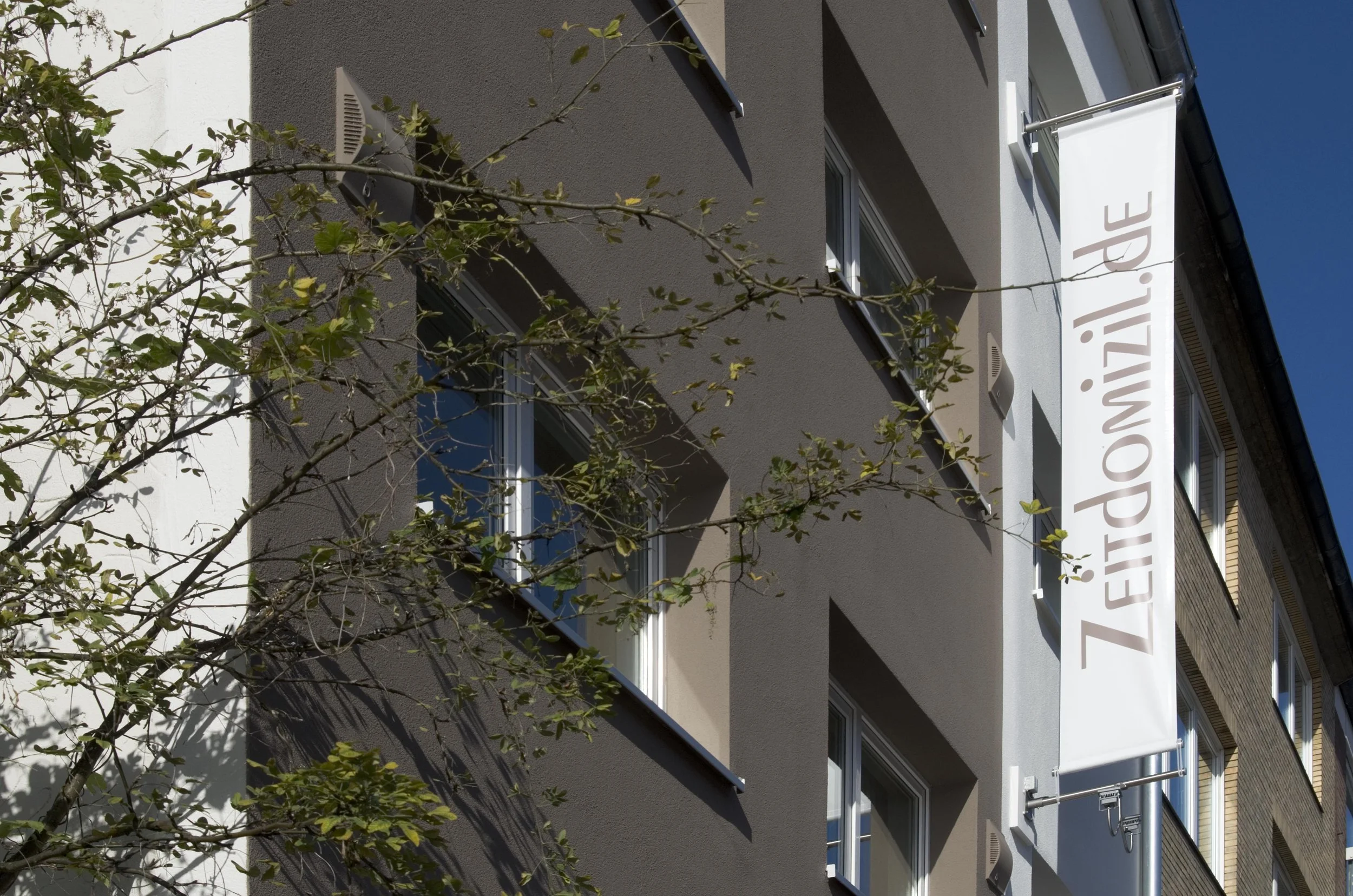 Ansicht eines modernen Mehrfamilienhauses mit Graufassade, mehreren Fenstern und einem weißen Banner mit der Aufschrift „ZEITOPTIQUE.PE“.