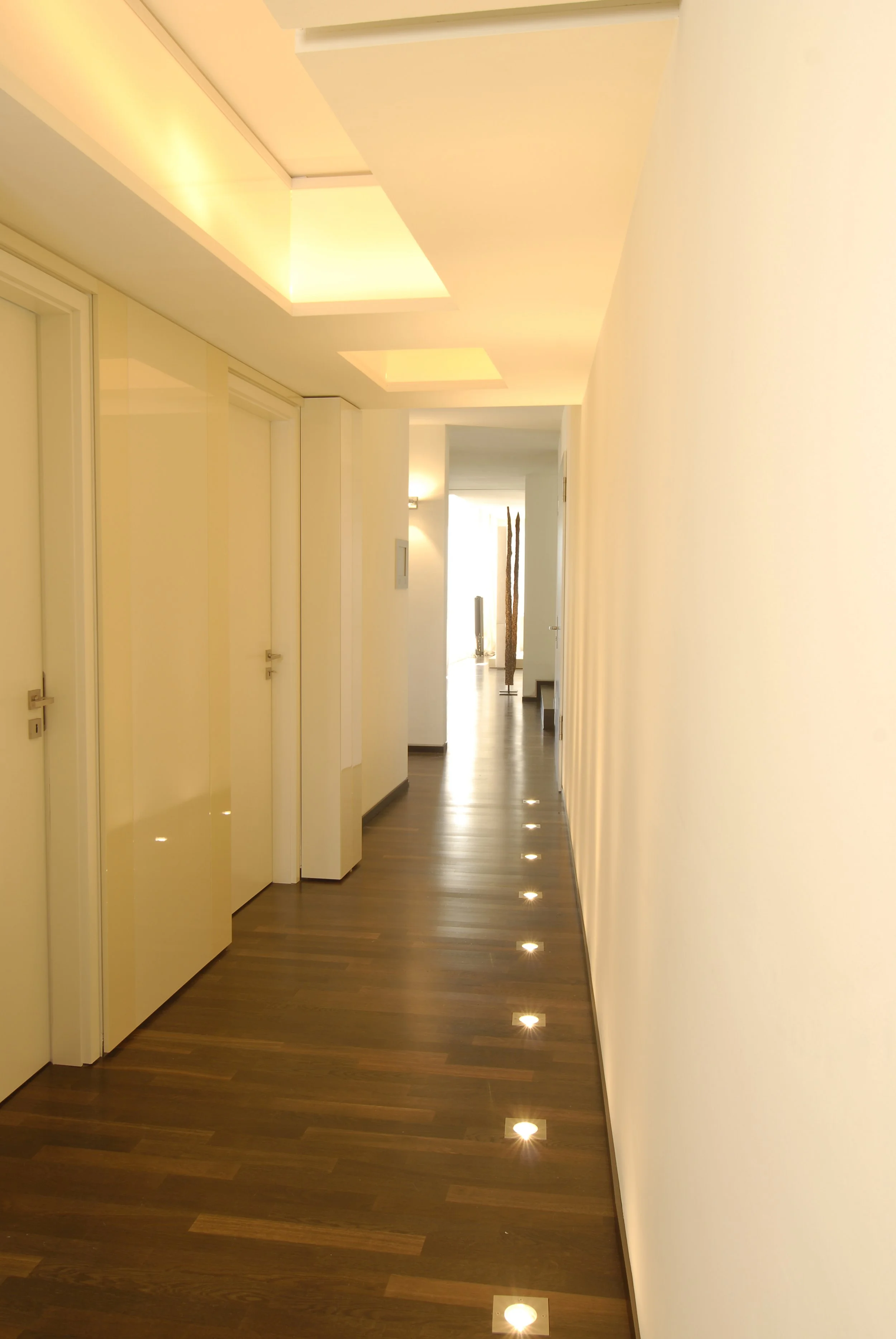 Ein heller, moderner Flur mit Holzfußboden, LED-Bodenleuchten und weißen Wänden. Im Hintergrund sind dekorative Skulpturen sichtbar.