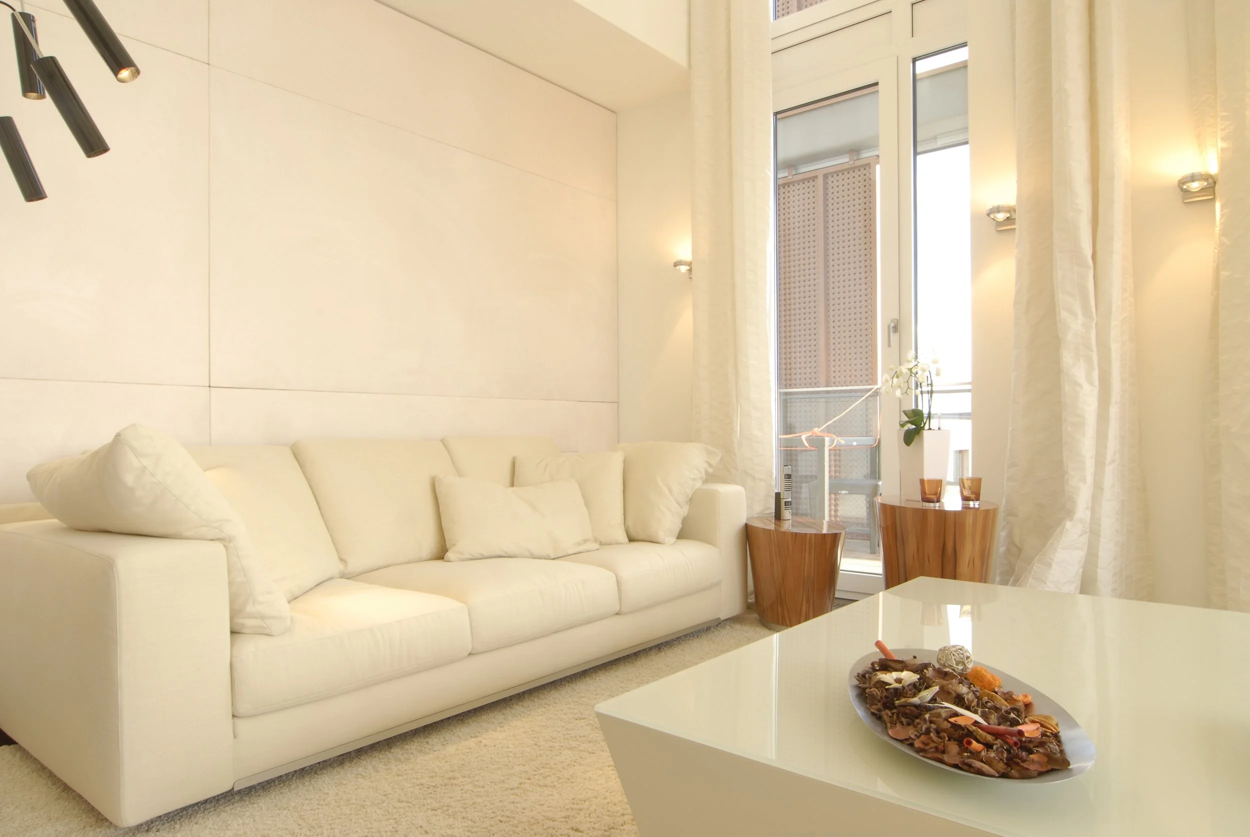 Heller Wohnzimmer mit cremefarbenem Sofa, zwei Holztischchen, große Fenster mit weißen Vorhängen, Wandlampen, Tisch mit Dekoration und Schale mit Süßigkeiten.