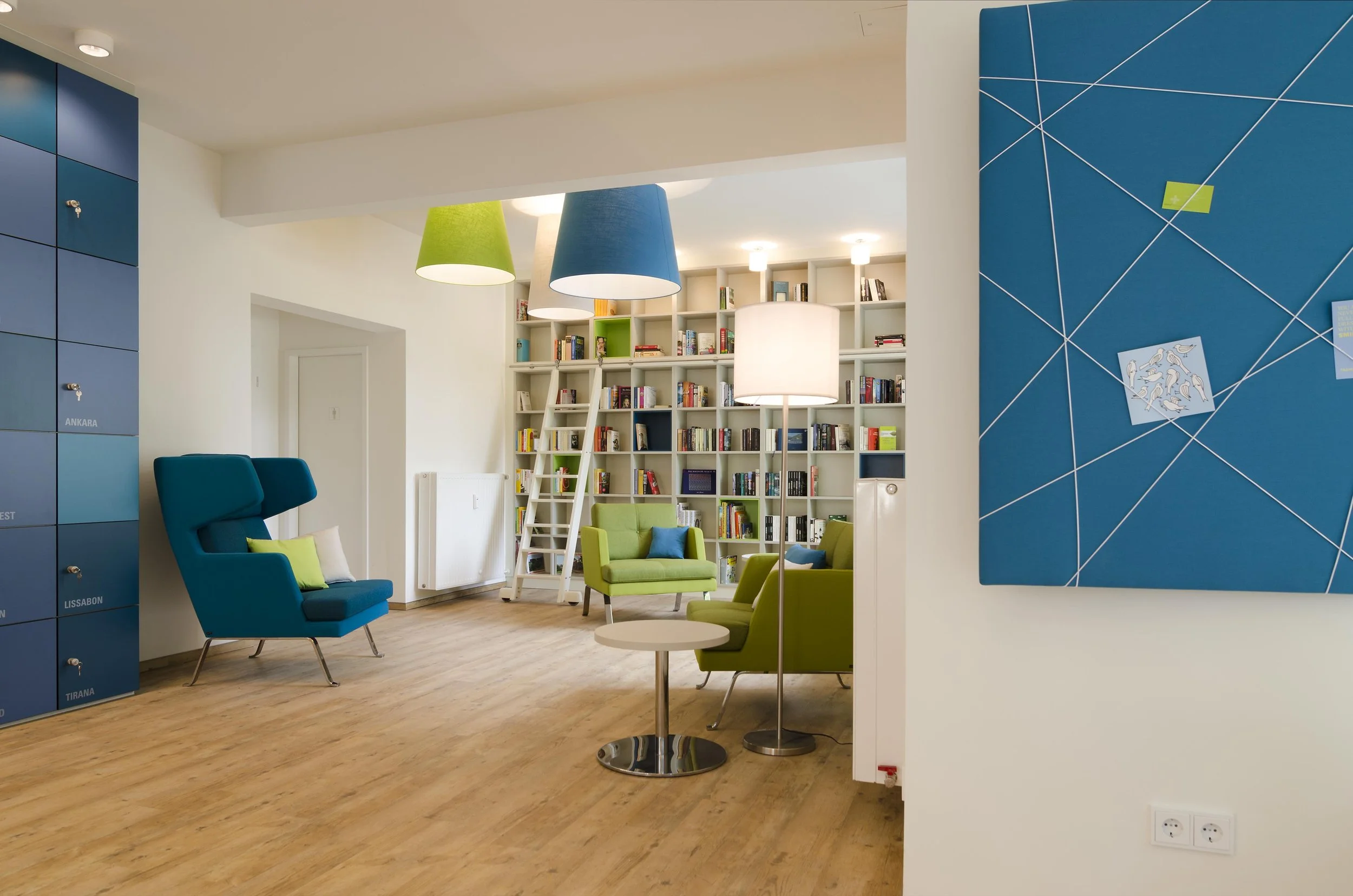 Gemütliche Leseecke mit grünen und blauen Sesseln, Bücherregale, Stehlampen und bunte Lampenschirme, moderne Einrichtung in einem Gemeinschaftsraum.
