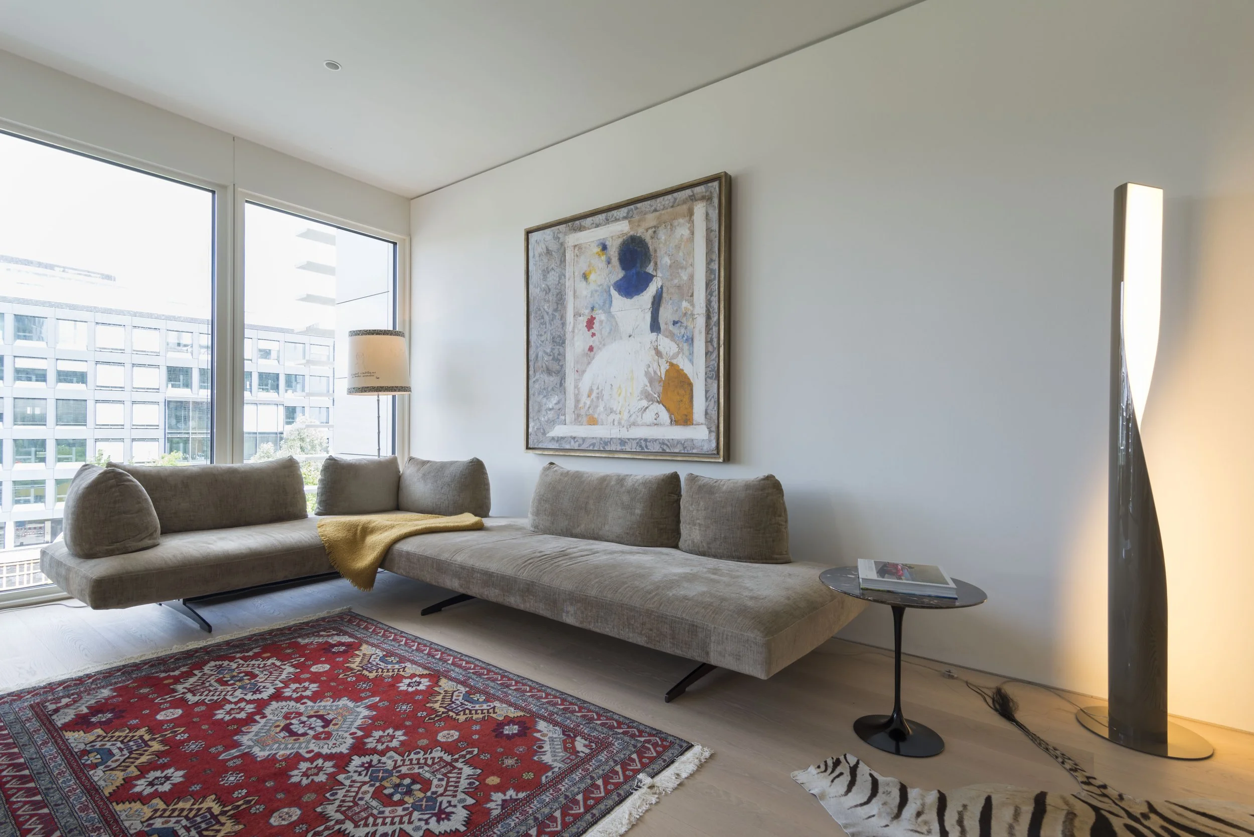 Ein modernes Wohnzimmer mit einem großen, hellen Sofa, einem bunten Teppich im roten Stil, einer stilvollen Stehlampe, einem kleinen Beistelltisch mit Zeitschriften und einem Zebra-Kopfteppich. Die Wand ist mit einem Kunstwerk dekoriert und große Fen