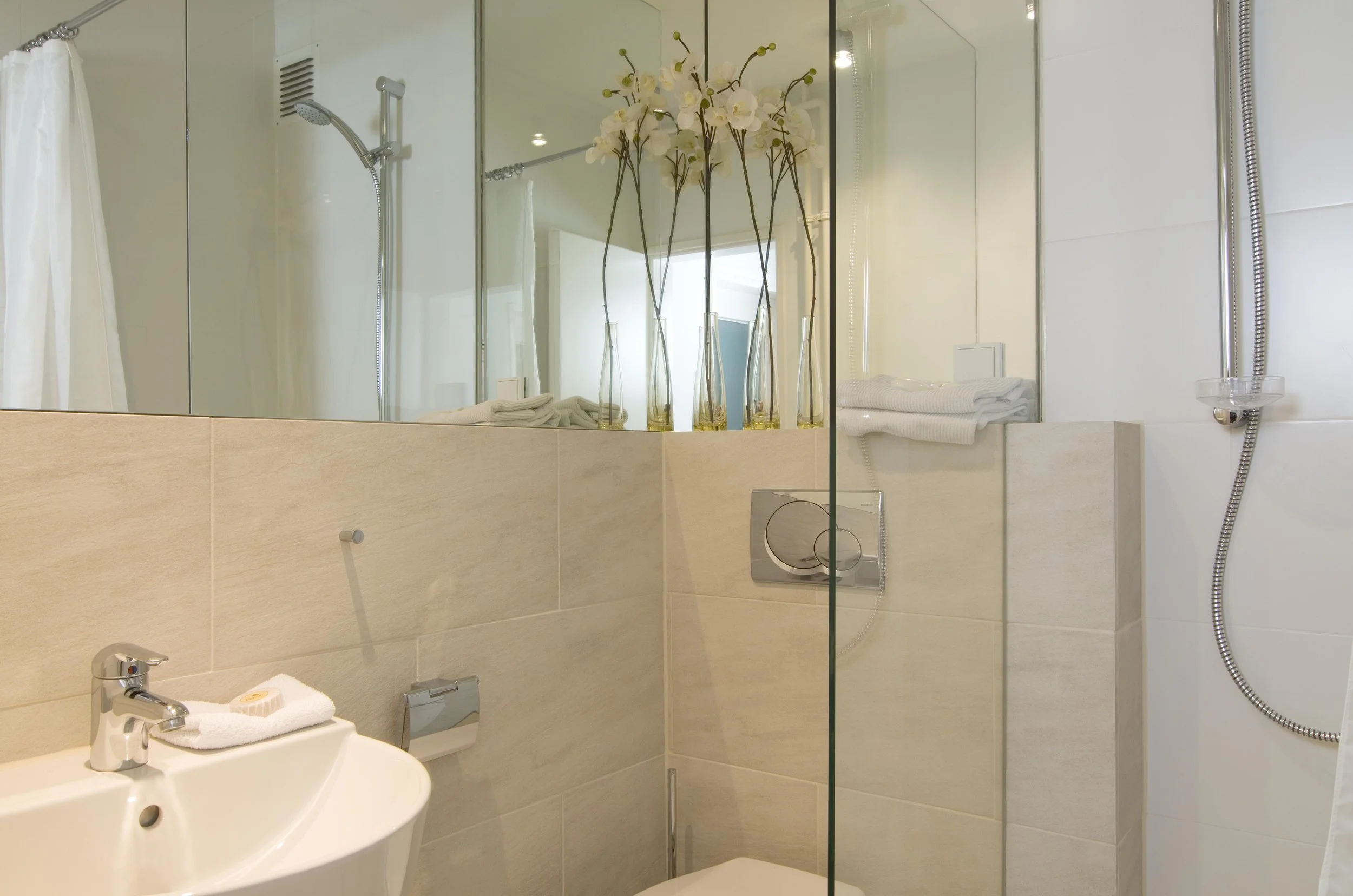 Modernes Badezimmer mit Dusche, Handwaschbecken, Spiegel, weißen Handtüchern und dekorativen weißen Blumen in Vasen.