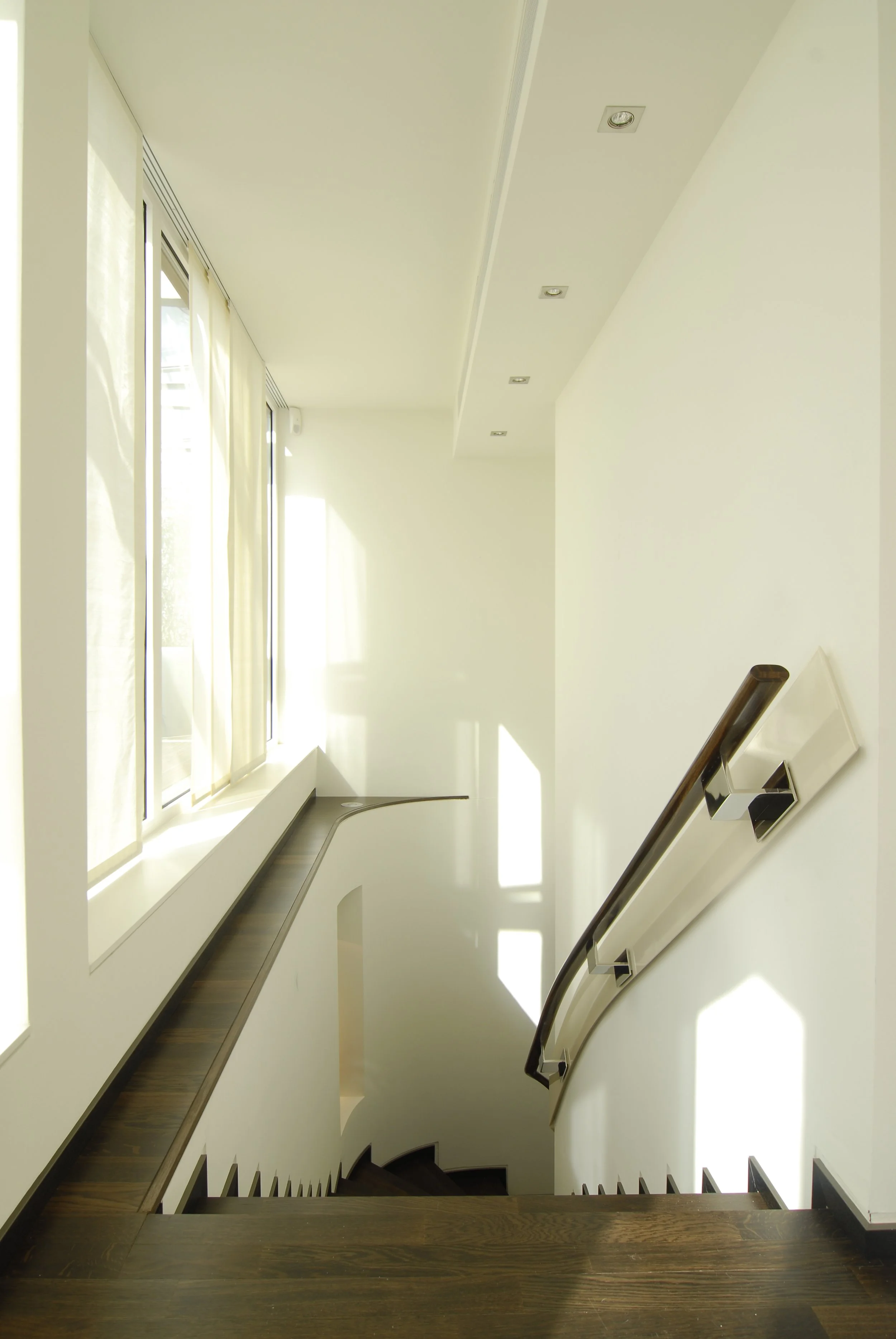 Innenansicht eines helle und modern gestalteten Treppenhauses mit Holztreppe, Handlauf, weißen Wänden und großen Fenstern mit Vorhängen, die Tageslicht einlassen.