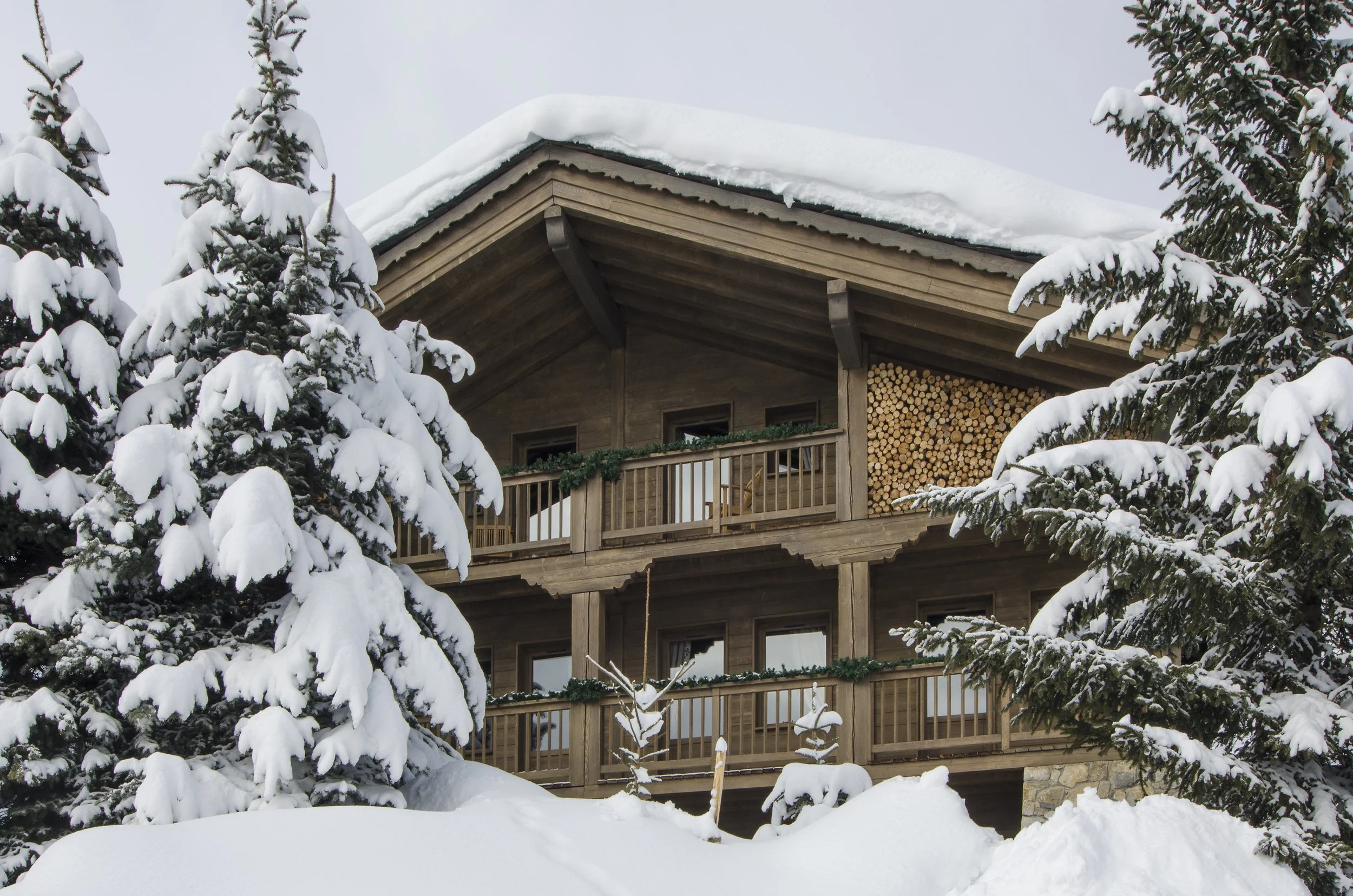 Holzhaus im Winter, umgeben von Schneebedeckten Tannenbäumen, mit Schneeschuttern auf dem Dach und auf den Bäumen.