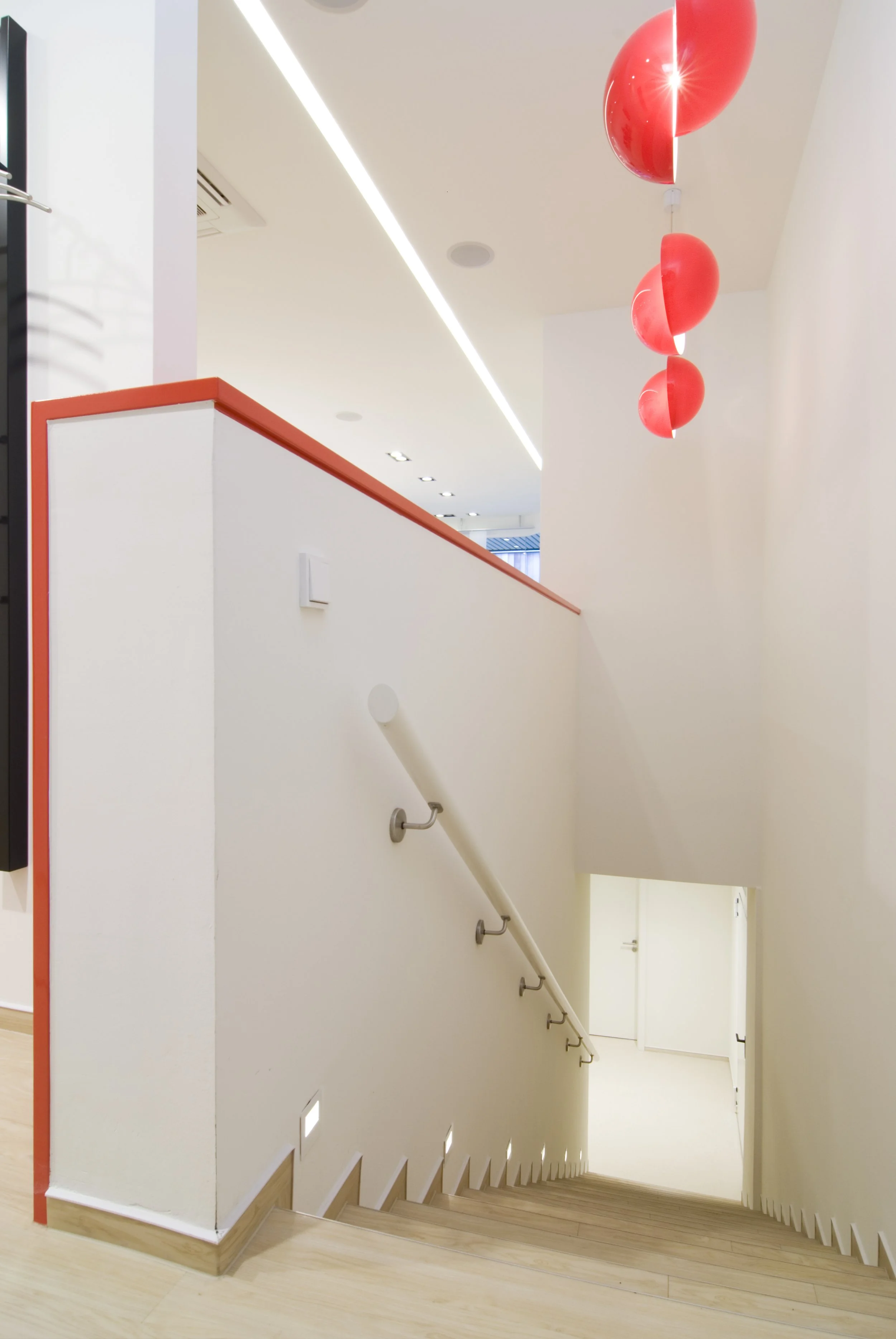 Weiße Treppe mit Holzstufen, Handlauf an einer weißen Wand, rote Lampen an der Decke, helle moderne Innenarchitektur.