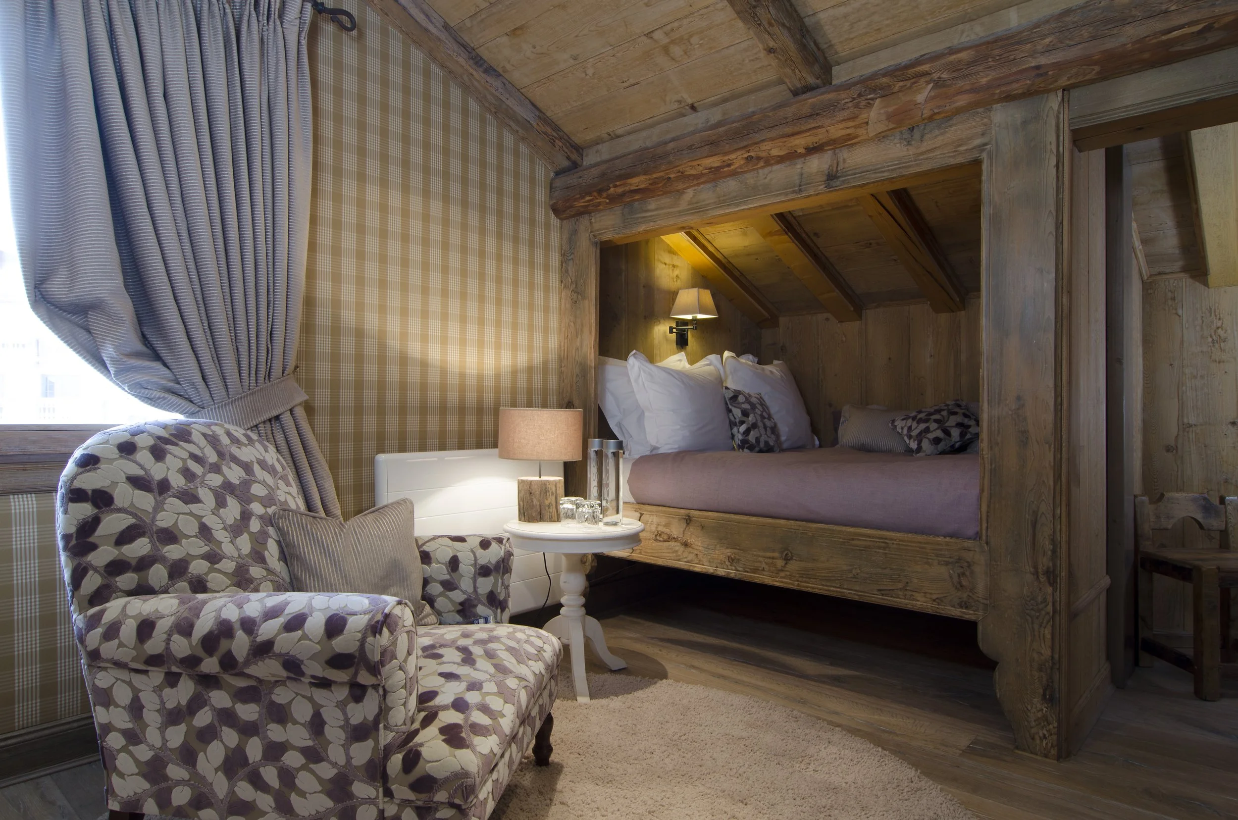 Gemütliches Schlafzimmer mit einer kleinen Sitzecke, einem Sessel mit Blumenmuster, einem kleinen weißen Tisch mit Lampe, und einem versteckten Bett im Holzofen, mit Kissen und Decke. Das Zimmer hat Holzwände und -decke, ein Fenster mit blauen Gardin