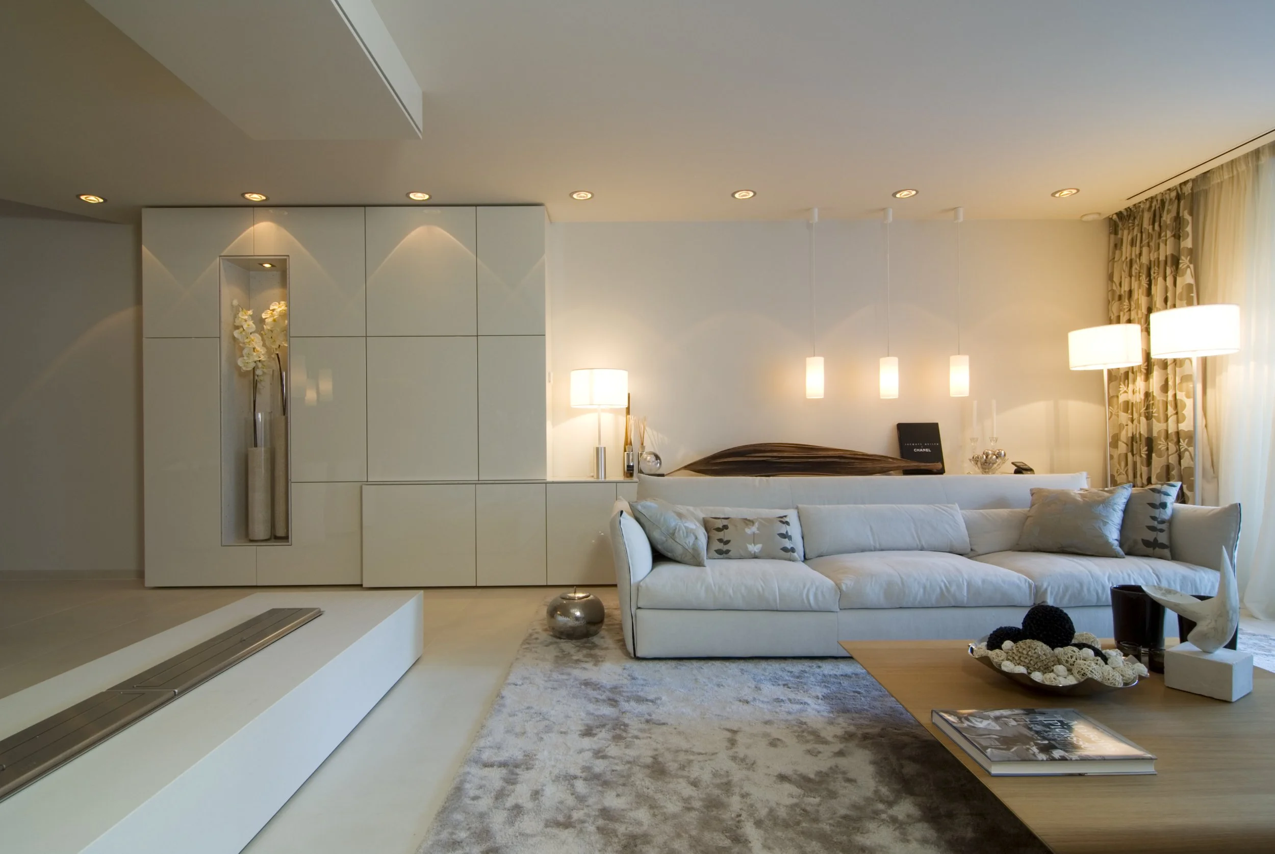 Modernes Wohnzimmer mit weißem Sofa, Stehlampen, hängenden Deckenleuchten, Couchtisch mit Dekorationen, Teppich und großen Fenstern mit Vorhängen.