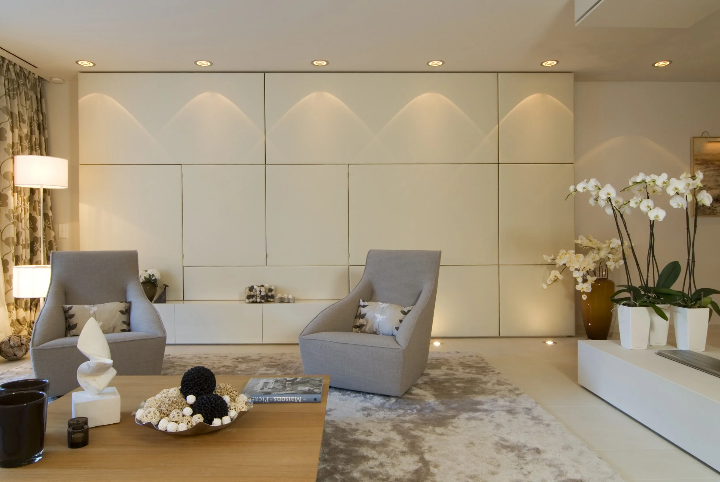 Moderner Wohnraum mit zwei grauen Sessel, weißen Orchideen in weißen Vasen, einem Couchtisch mit Dekorationen und einem großen weißen Schrank im Hintergrund.
