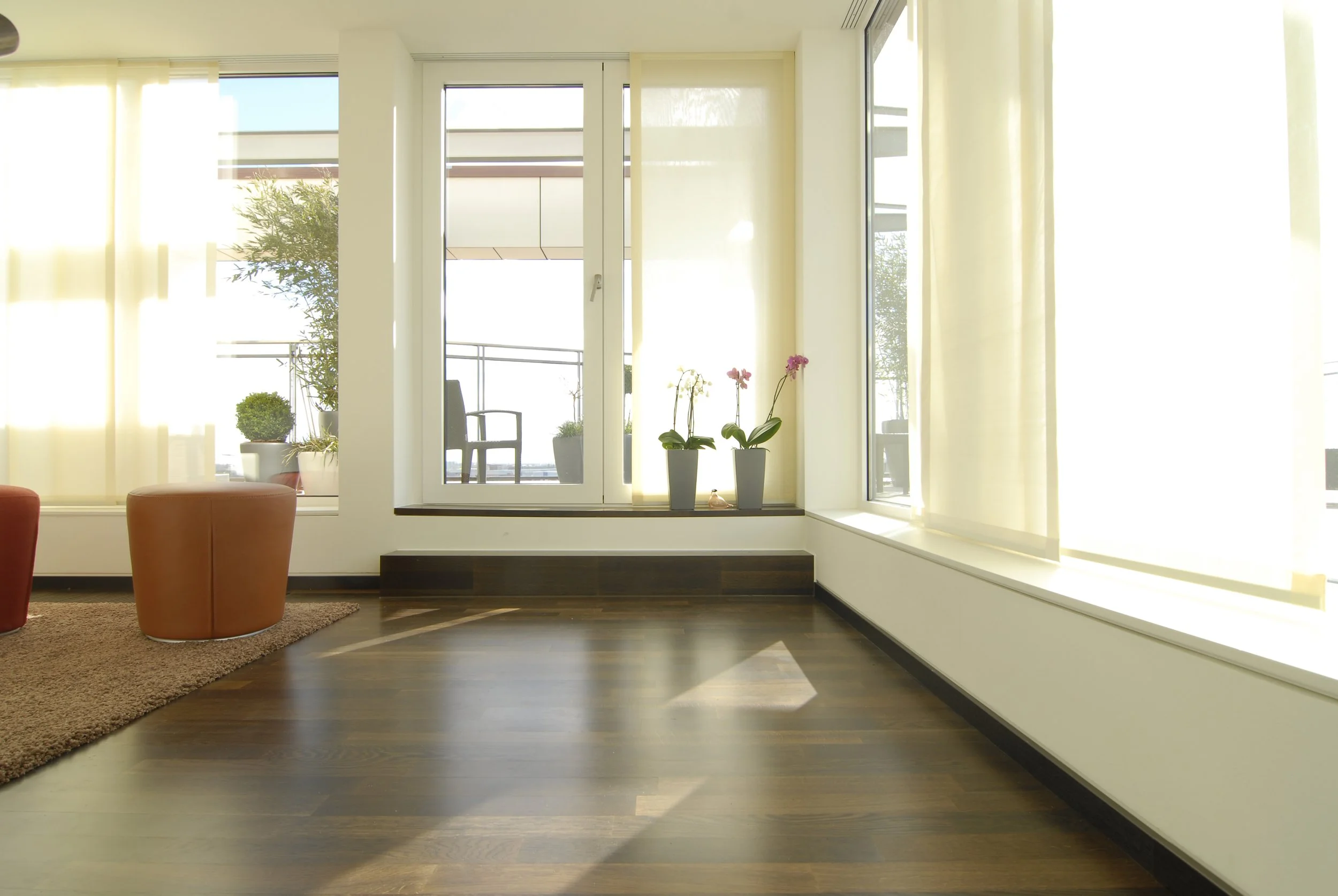 Innenraum mit Holzboden, großen Fenstern mit durchsichtigen Vorhängen, Pflanzkübeln mit Orchideen, einem Balkon mit Stuhl im Hintergrund.