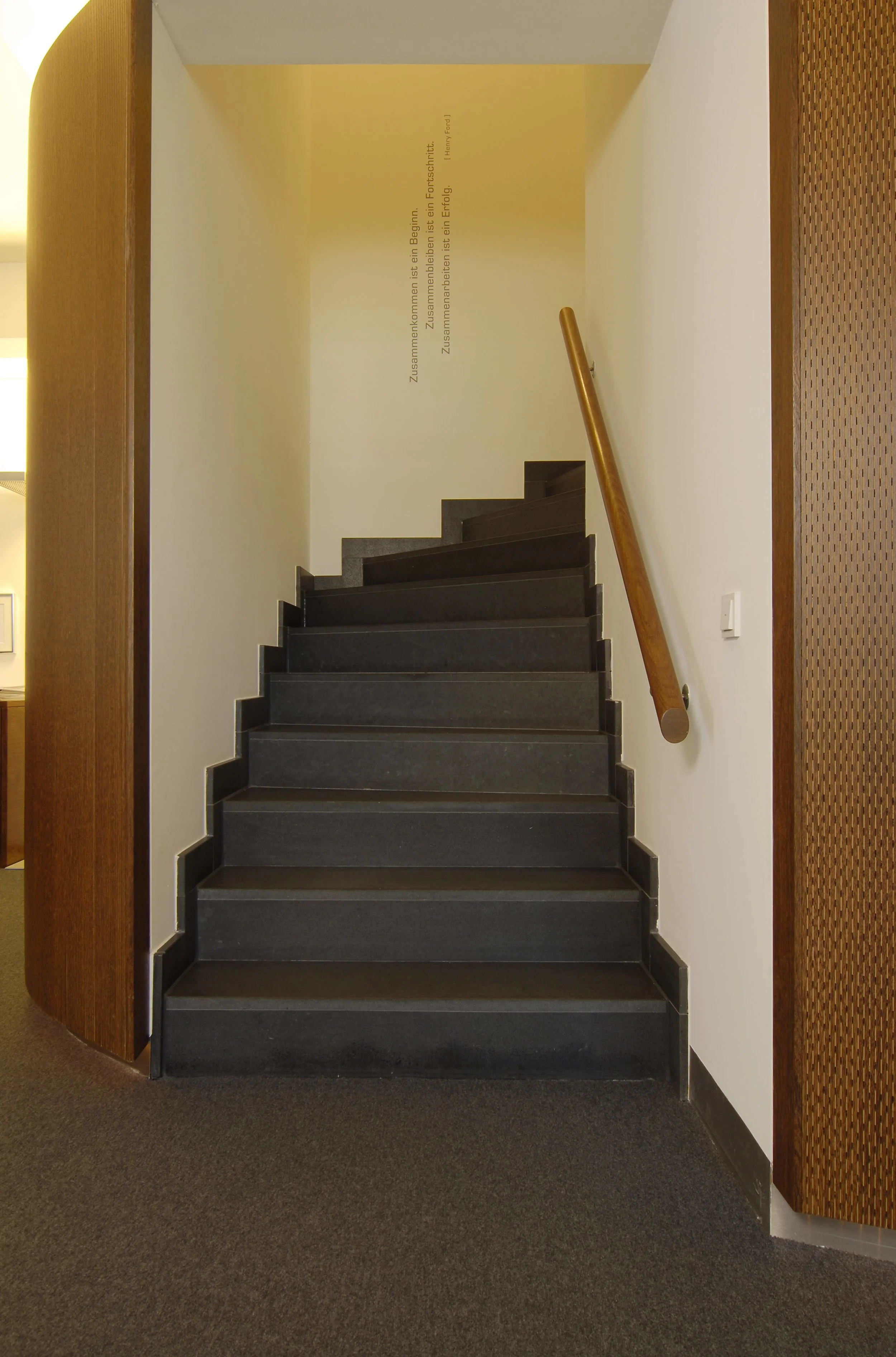 Schwarze Treppe mit Holzhandlauf in einem Innenraum, Wand mit einem Zitat in deutscher Sprache, an der Decke Lichtquellen, Teppichboden