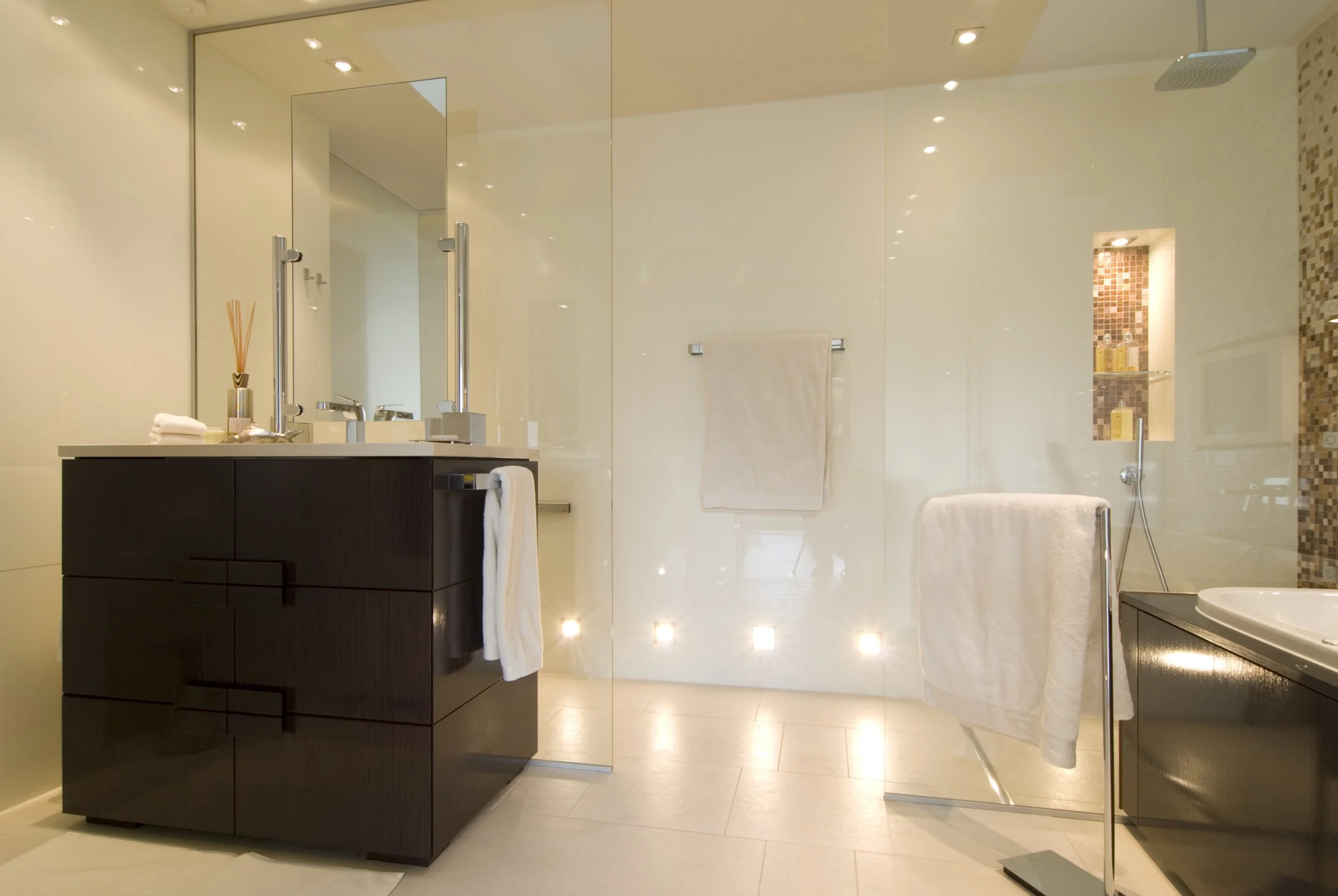 Moderner Badezimmer mit dunklem Waschtisch, großen Spiegel, Glasdusche mit Mosaikverkleidung, Handtuchheizung und Rollenhandtüchern.