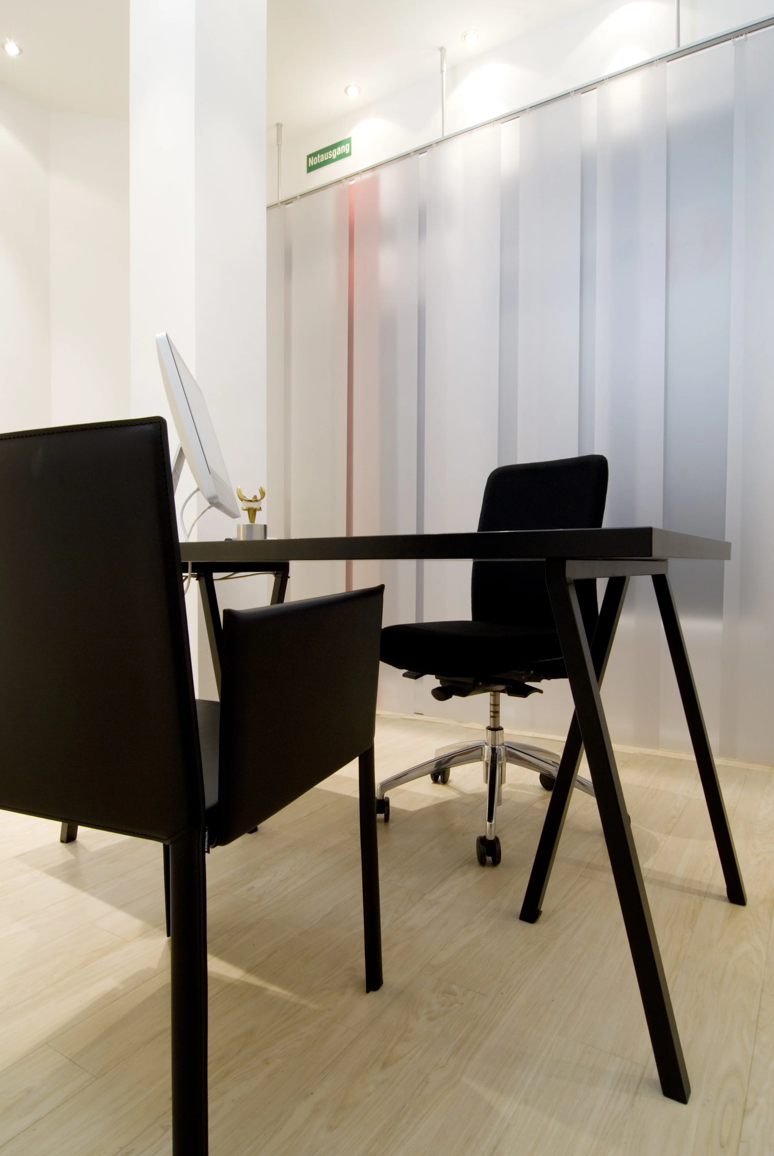Ein leeres Büro mit einem schwarzen Schreibtisch, schwarzen Stühlen und einem weißen Computer. Es gibt eine Notausgangstafel oben an der Wand.