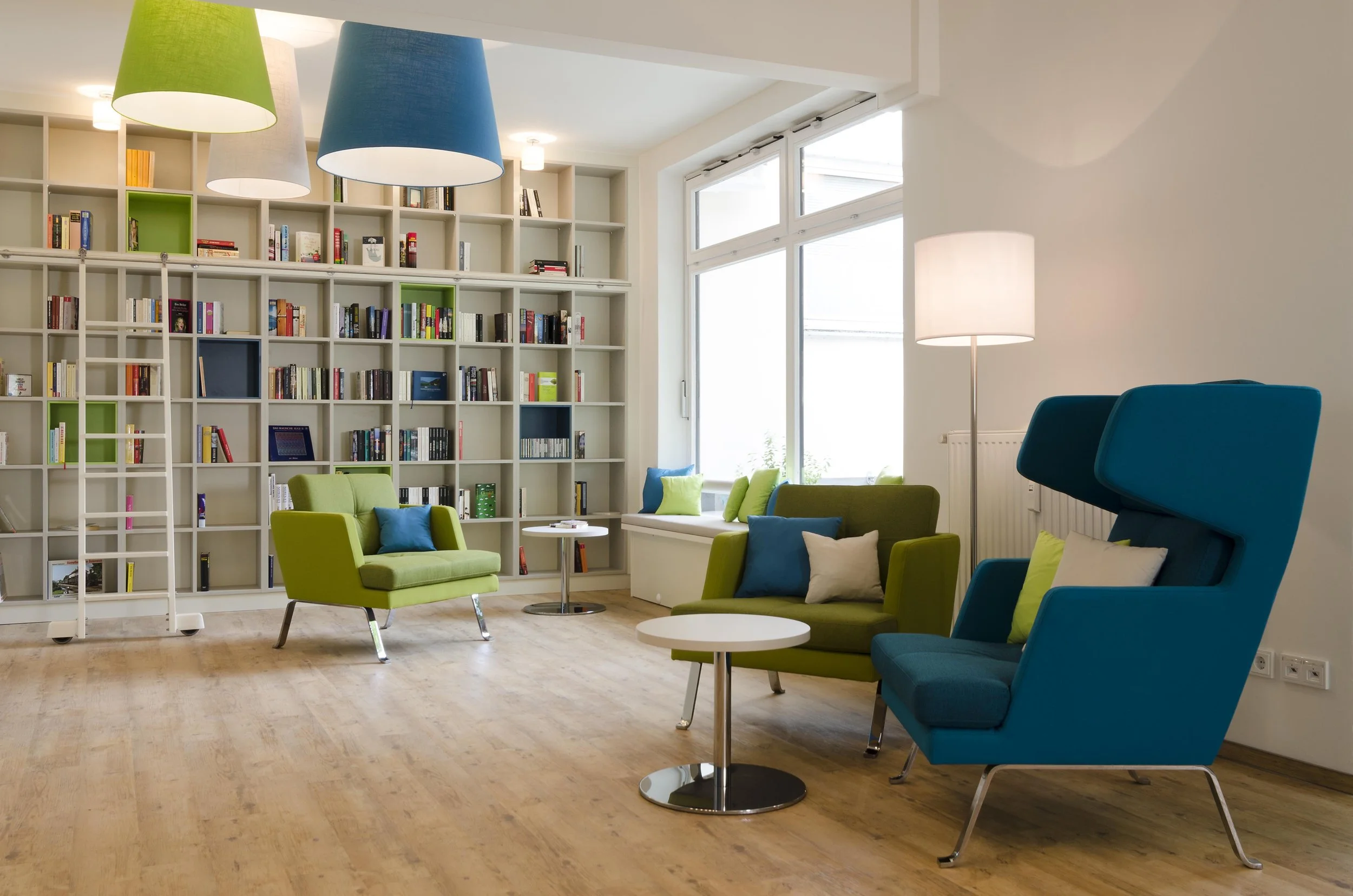 Gemütliches Lese- und Loungebereich mit bunten Sesseln, einem Bücherregal, großem Fenster und Stehlampen.
