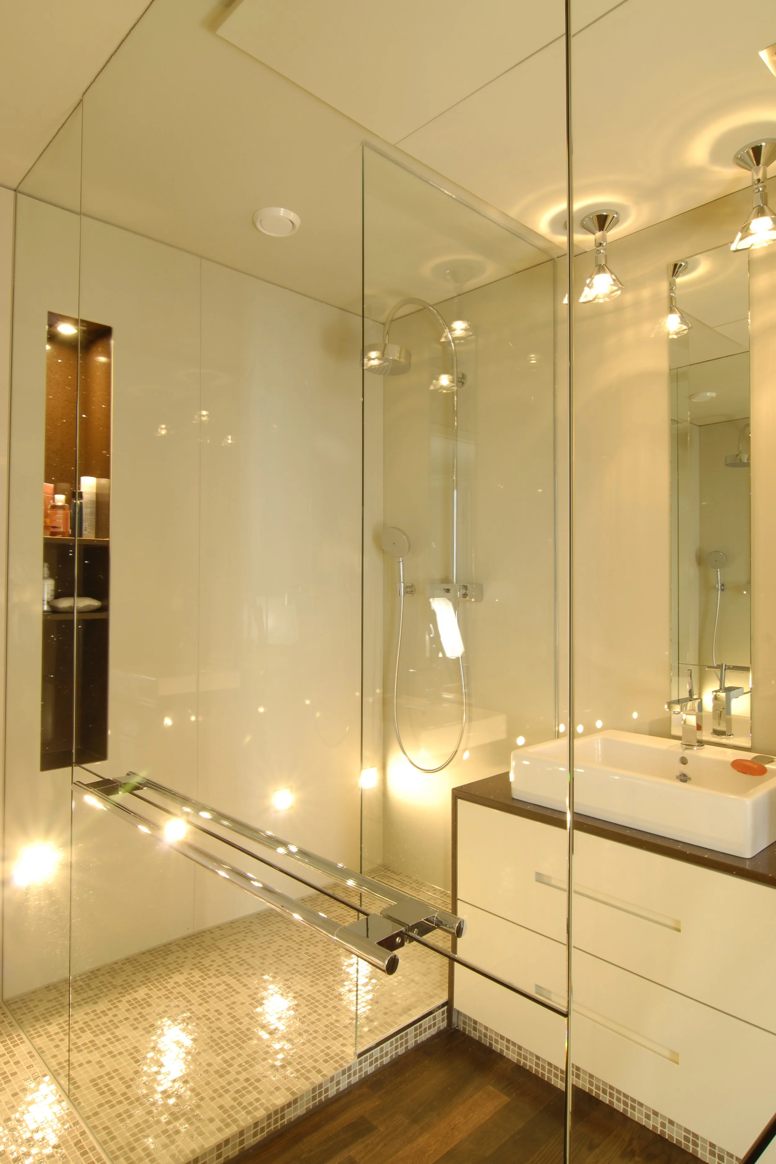Moderne Badezimmer mit begehbarer Dusche, großer Spiegel, weißem Waschbecken und dunklem Holzfußboden, warmes Licht.