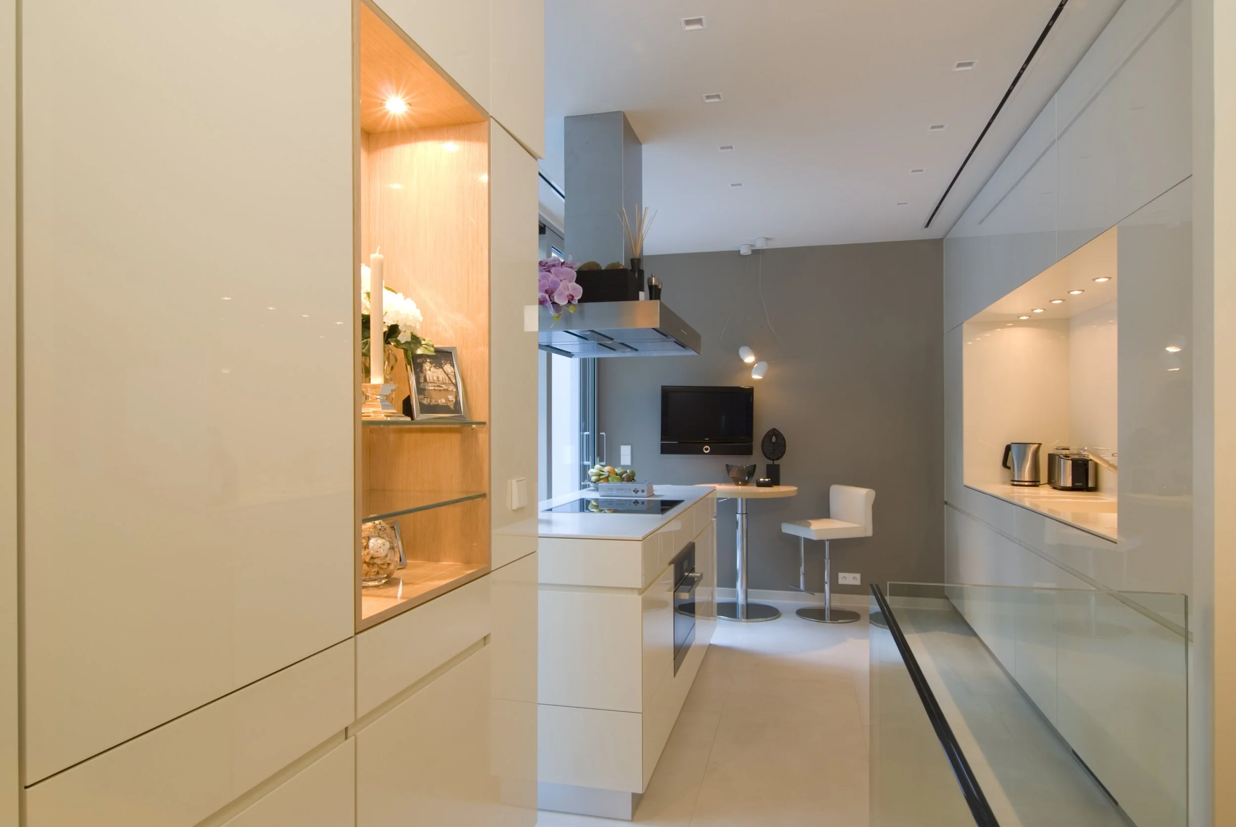 Moderne, minimalistische Küche mit weißen Schränken, einer kleinen Theke mit Barhockern, einem Fernseher an der Wand und Küchenutensilien auf der Ablage.