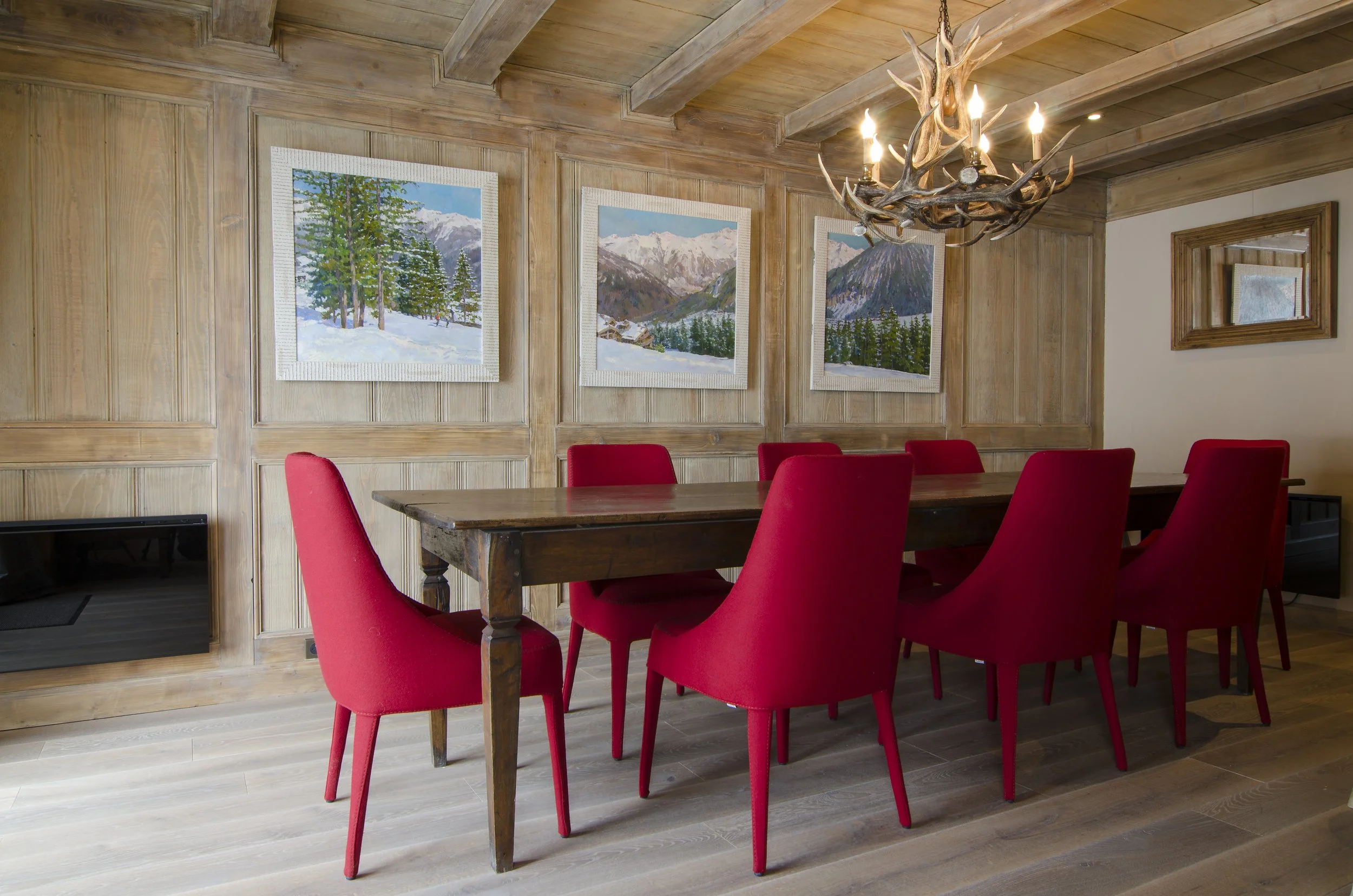 Ein Esszimmer mit einer großen Tafel, roten Stühlen, einer antler Kronleuchter und Bildern von Berglandschaften an der Wand.