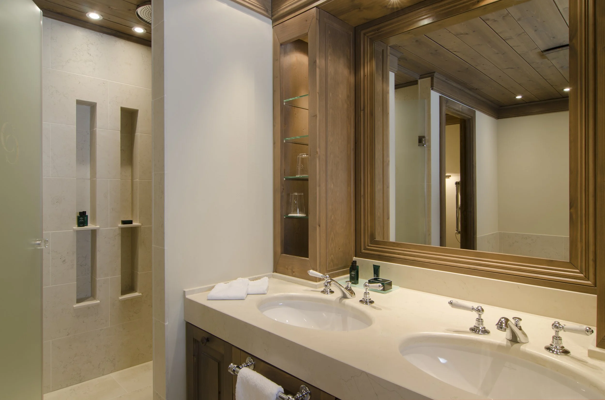 Badezimmer mit Doppelwaschbecken, großem Spiegel, Holzdekor, Duschecke mit Nischen, Handtüchern und Pflegeprodukten.