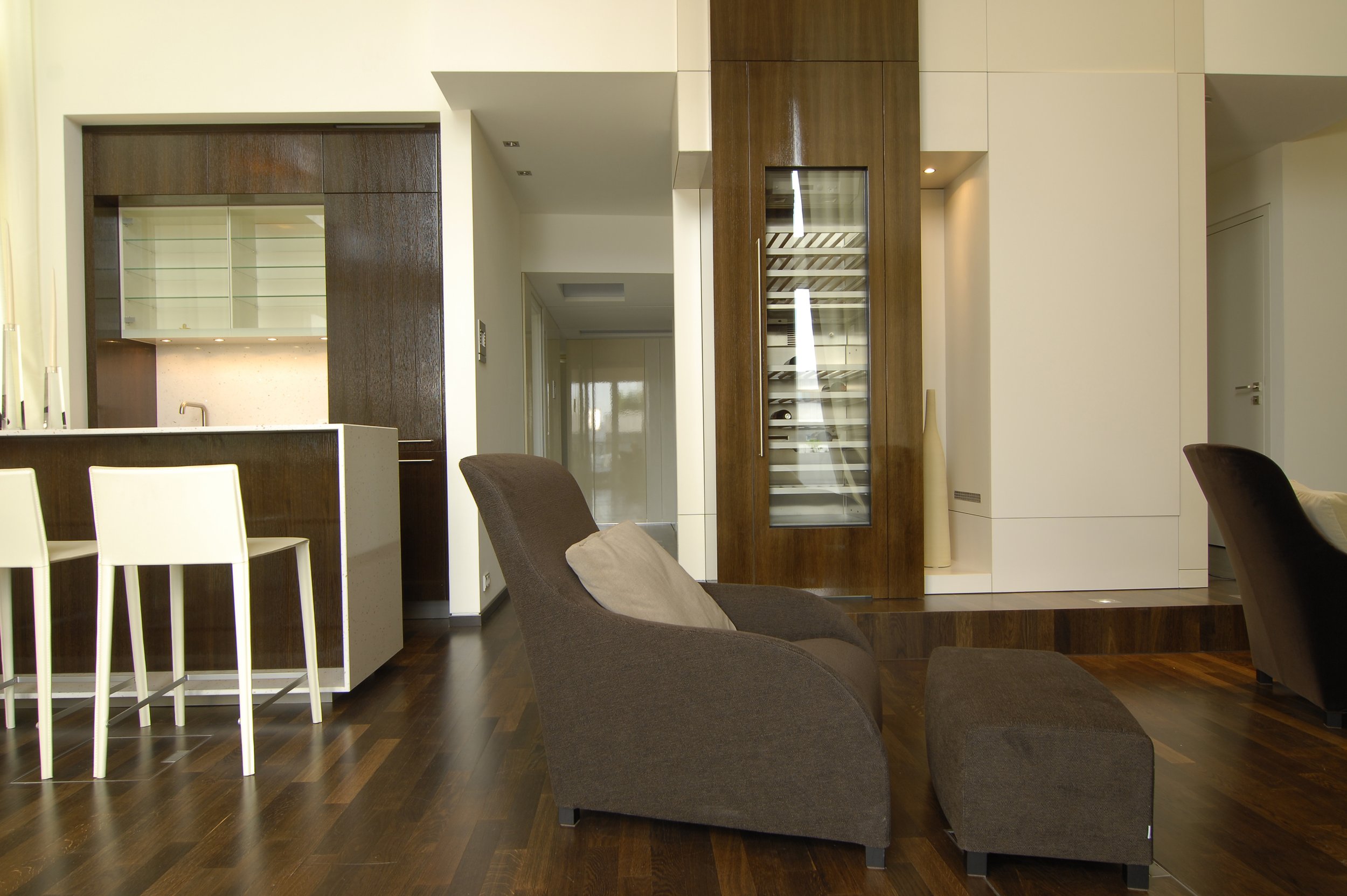 Moderner, minimalistisch eingerichteter Wohnraum mit braunen Sesseln, einem dunklen Holztisch, weißen und braunen Küchenmöbeln sowie einem Einbauschrank mit Schlafsystem im Hintergrund.
