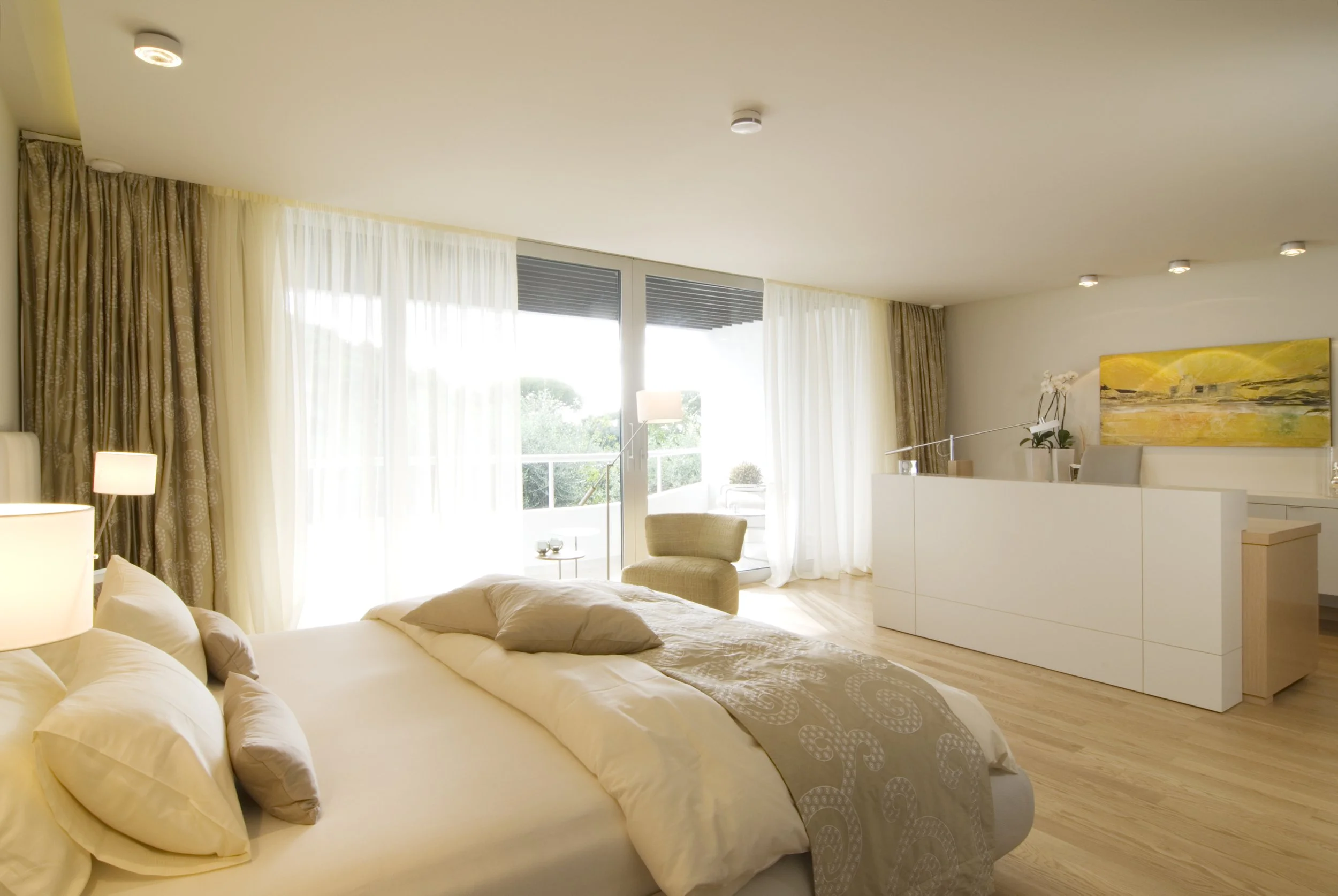 Ein modernes Schlafzimmer mit großen Fenstern, weißen Vorhängen, einem Bett mit weißen und beigen Bettwäsche, einem Sessel auf der Terrasse, einer weißen Kommode und einem bunten Gemälde an der Wand.