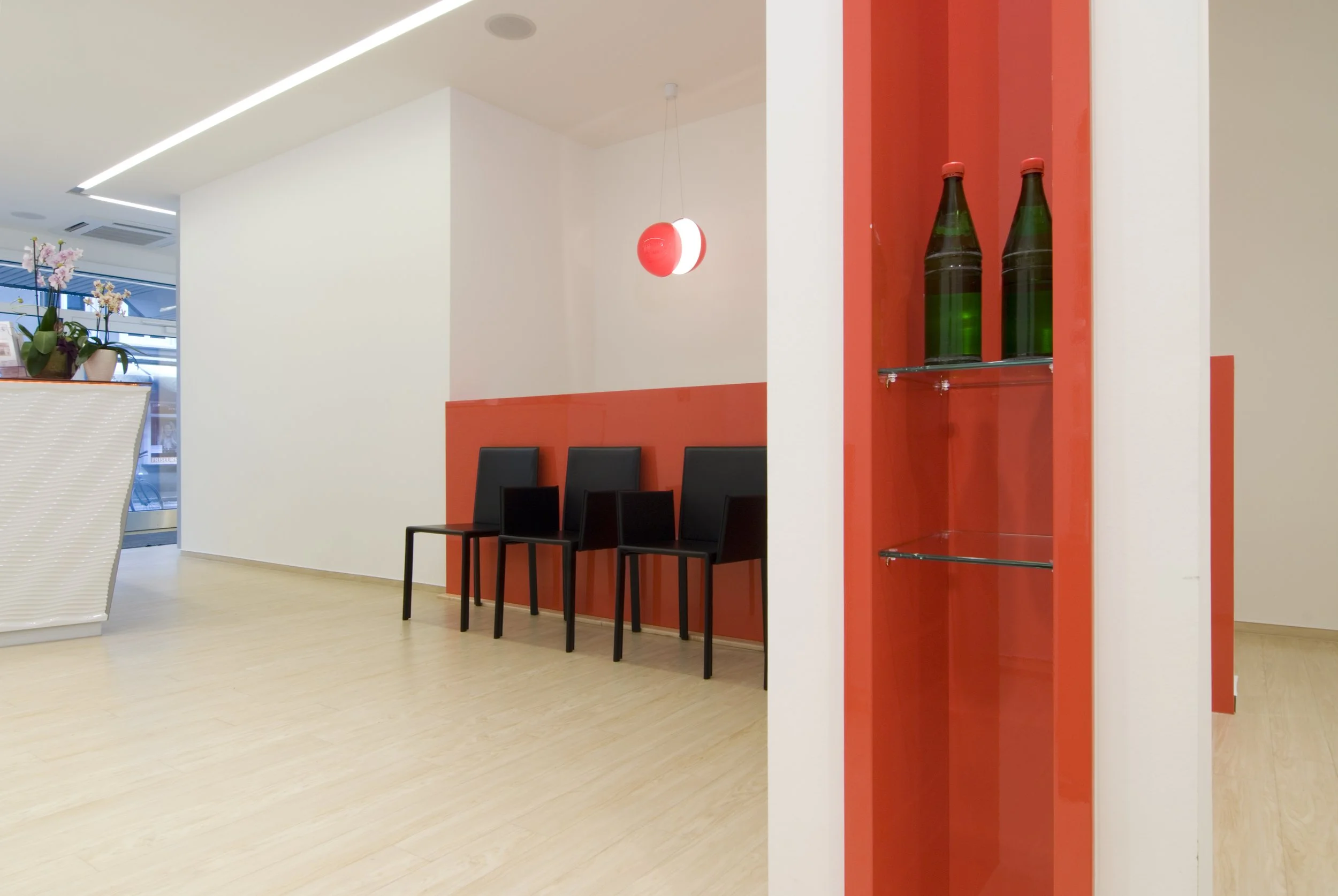 Ein modernes Wartezimmer mit drei schwarzen Stühlen, einer roten Wand mit Beleuchtung und dekorierten Flaschen auf einem Glasregal.