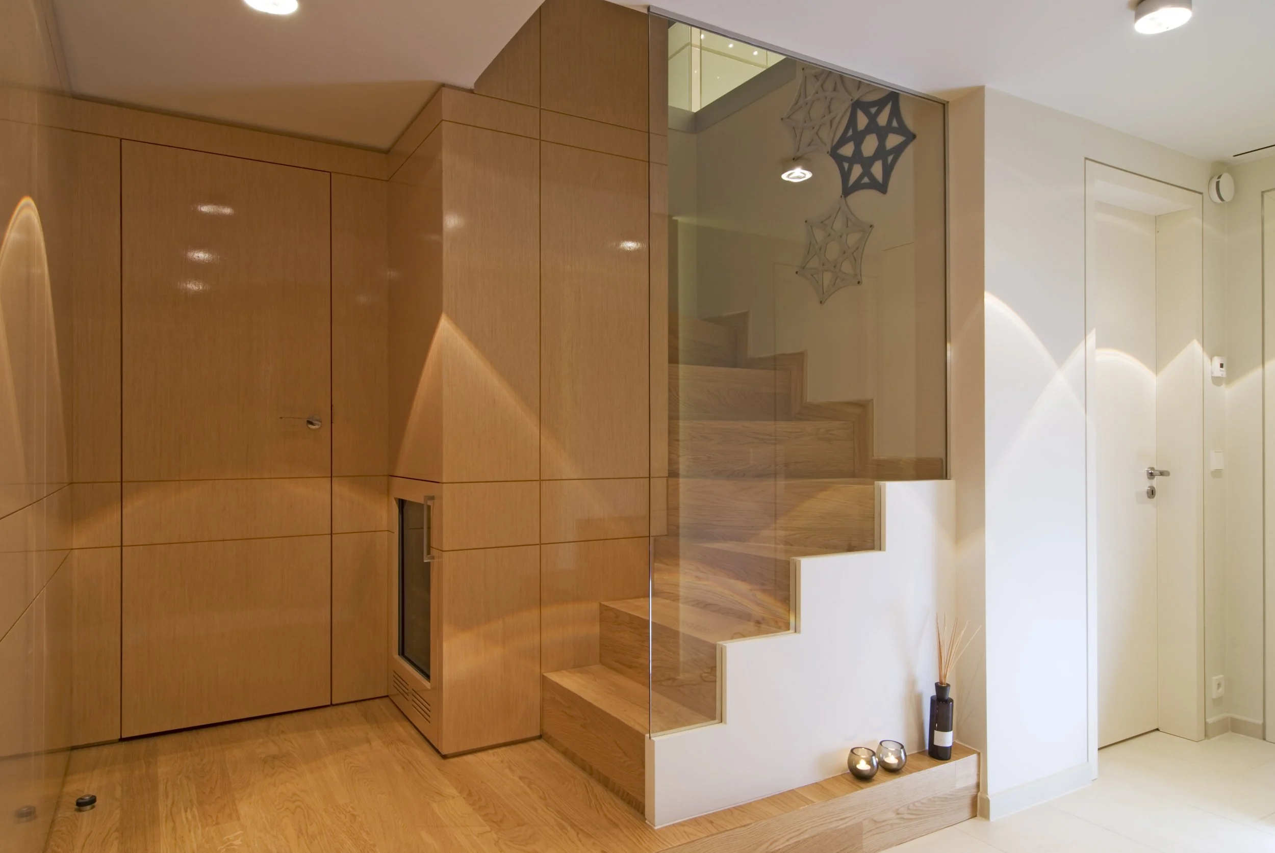 Innenaufnahme eines modernen Hauses mit Holztreppe, Holzverkleidung, Deko-Elementen wie Kerzen und Steingefäßen, und gehobener Beleuchtung.