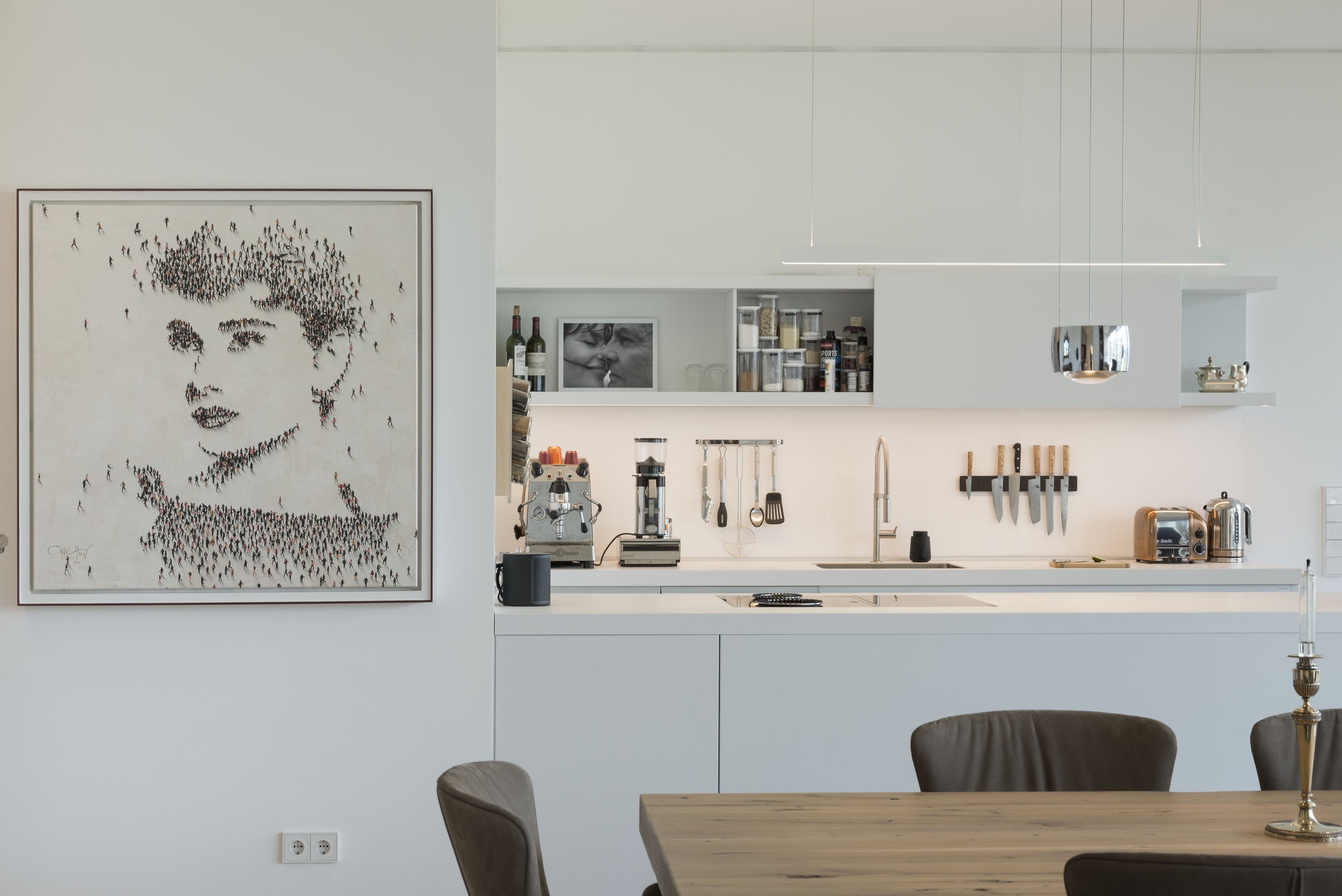 Moderne white Küchenzeile mit Kochfeld, Spüle, Messerhalter mit Messern, Toaster, Kaffeemaschine und Regalen mit Gewürzen. Küchenwand mit zwei Fotos, einer Kaffeemaschine und einer Zuckerdose. Im Vordergrund ein Holztisch mit Stühlen und eine Kerze. 