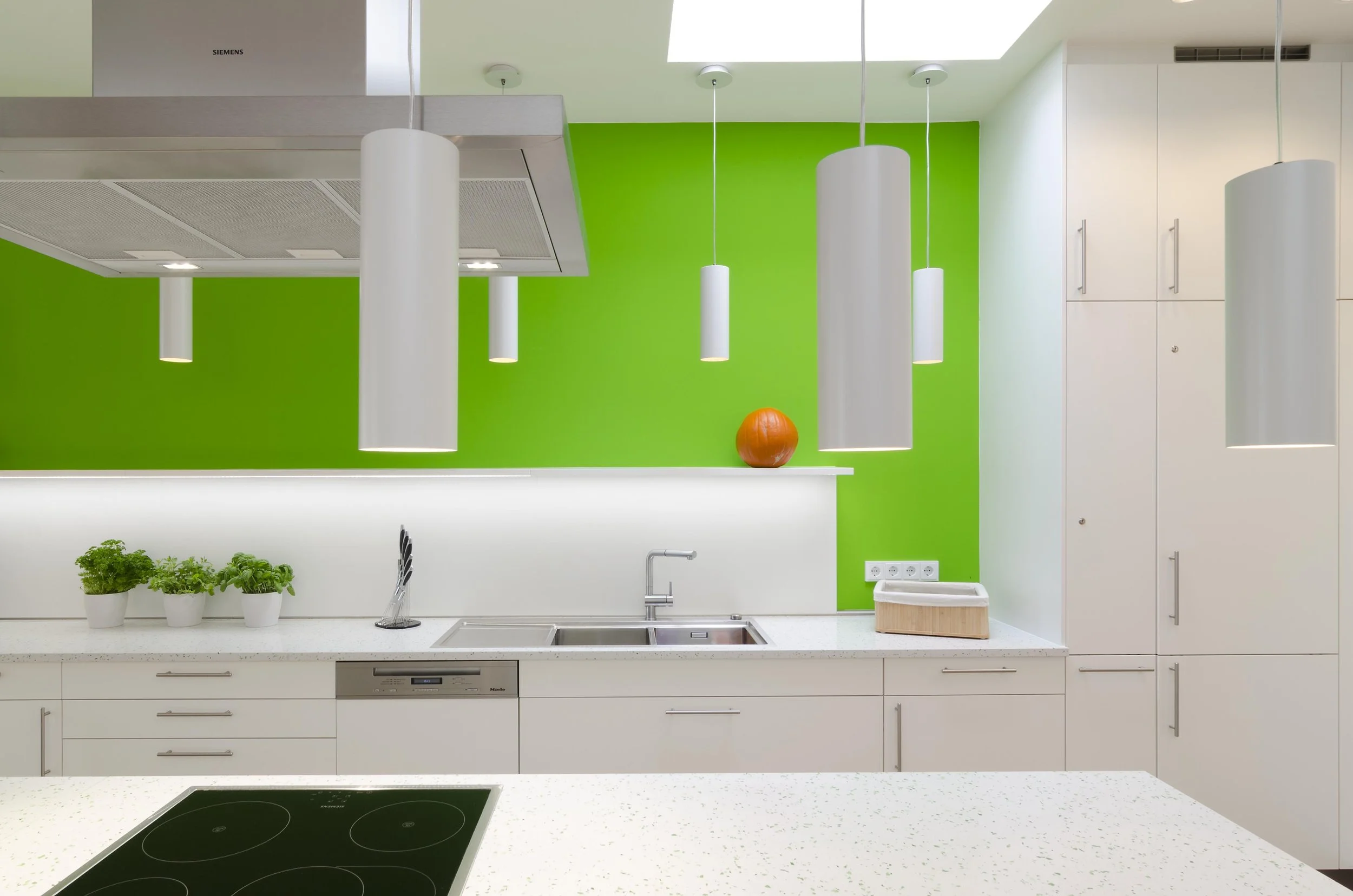 Moderne Küche mit weißen Schränken, grüner Wand, vier weißen Pendelleuchten, Herd, Spüle, Küchenutensilien, Topfpflanzen und einer orangefarbenen Kugel auf der Theke.