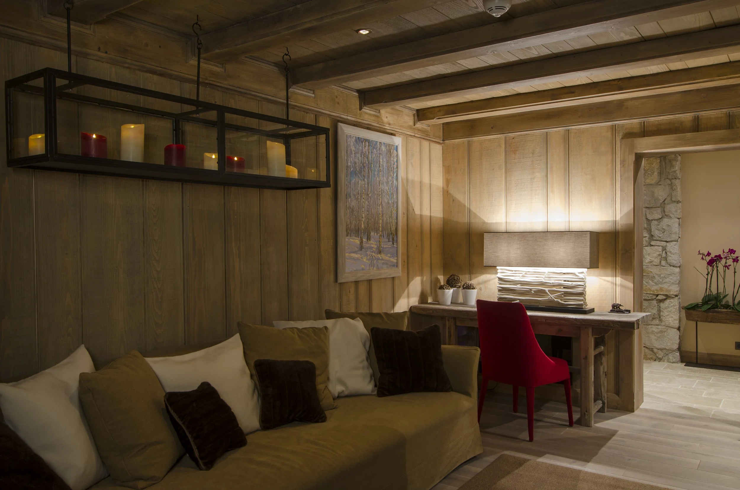 Gemütliches Wohnzimmer mit Holzvertäfelung, einer Couch mit Kissen, einem Schreibtisch mit rotem Stuhl, Wandbilder und Kerzen.