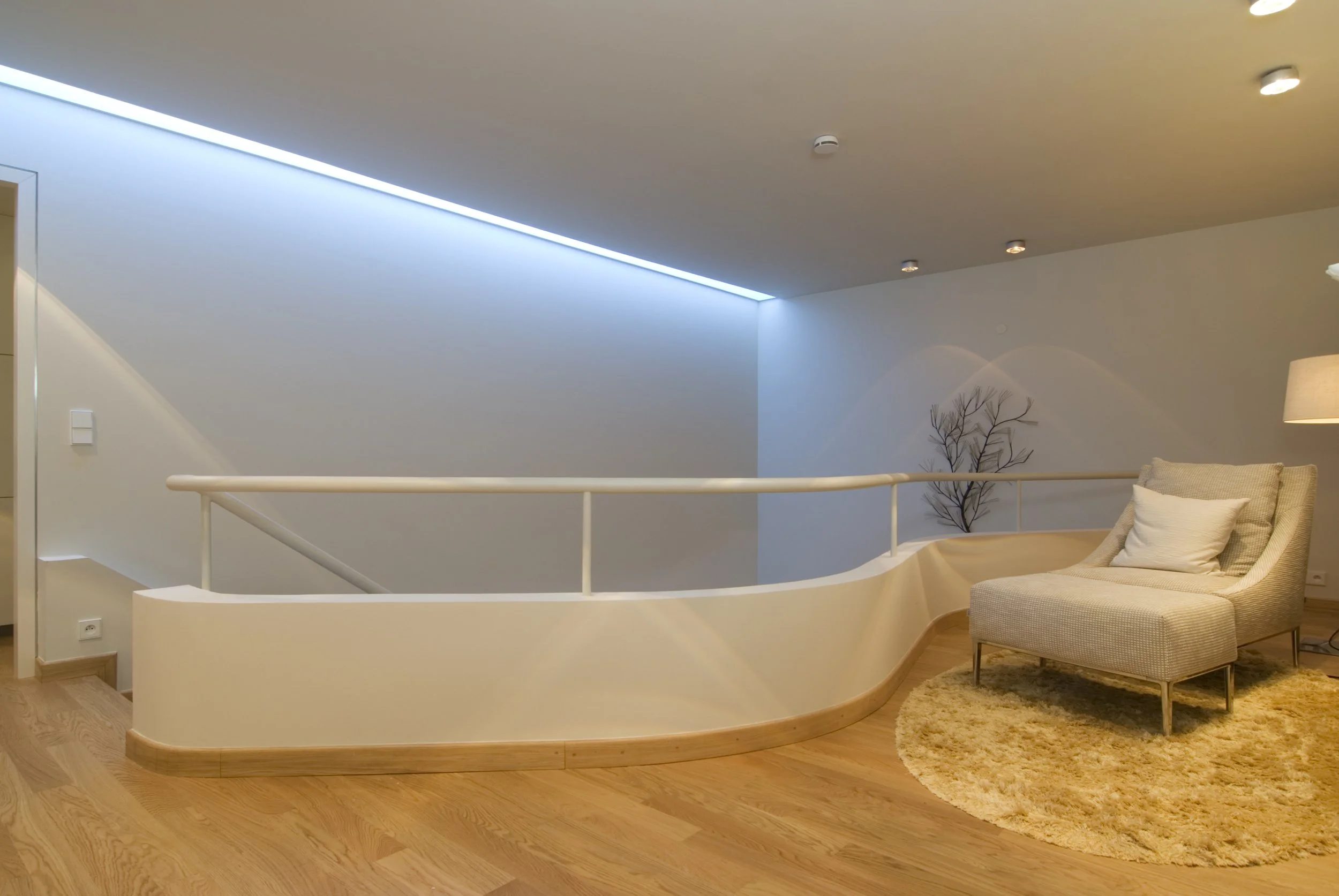 Helles Wohnzimmer mit Holzfußboden, cremefarbenem Sessel, hellem Teppich, Stehlampe, Wandbild eines Baumes, abgetrennter Bereich durch weiße, geschwungene Wand und Geländer, Deckenbeleuchtung, blaulichtdurchfluteter Raum.