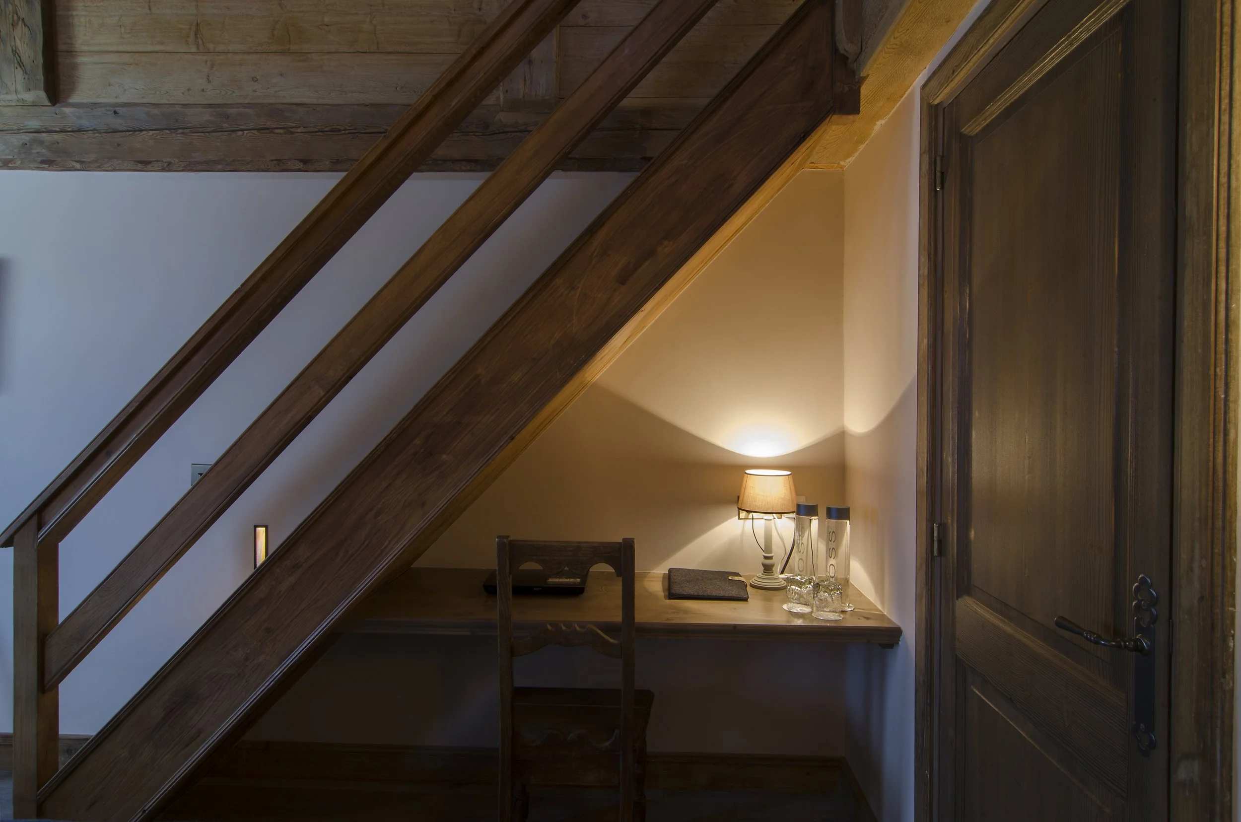 Ein kleiner Schreibtisch unter einer Treppe, mit Stehlampe, Wasserflaschen, Gläsern und einem Notizbuch, in einem warm beleuchteten Raum.