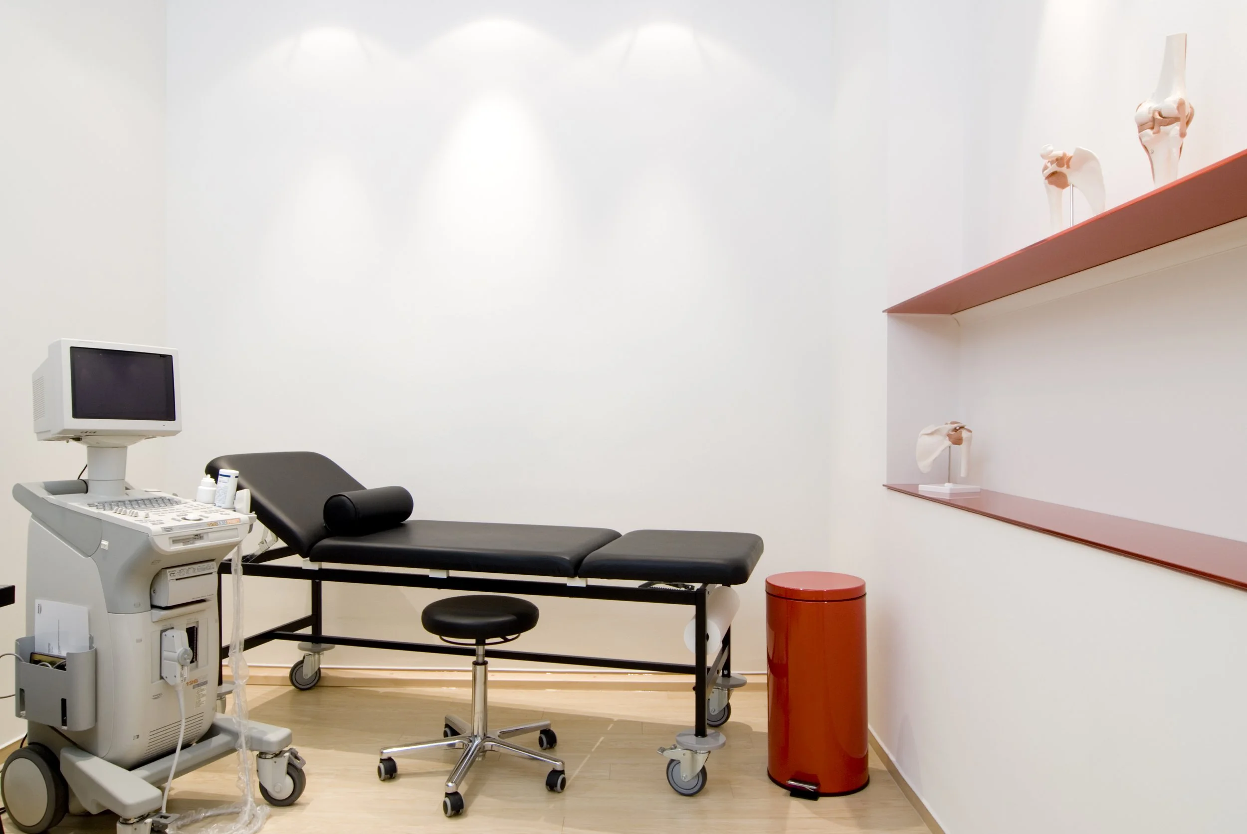 Ärztezimmer mit Untersuchungsliege, Ultraschallgerät, Stuhl, Mülleimer und anatomische Modelle an der Wand