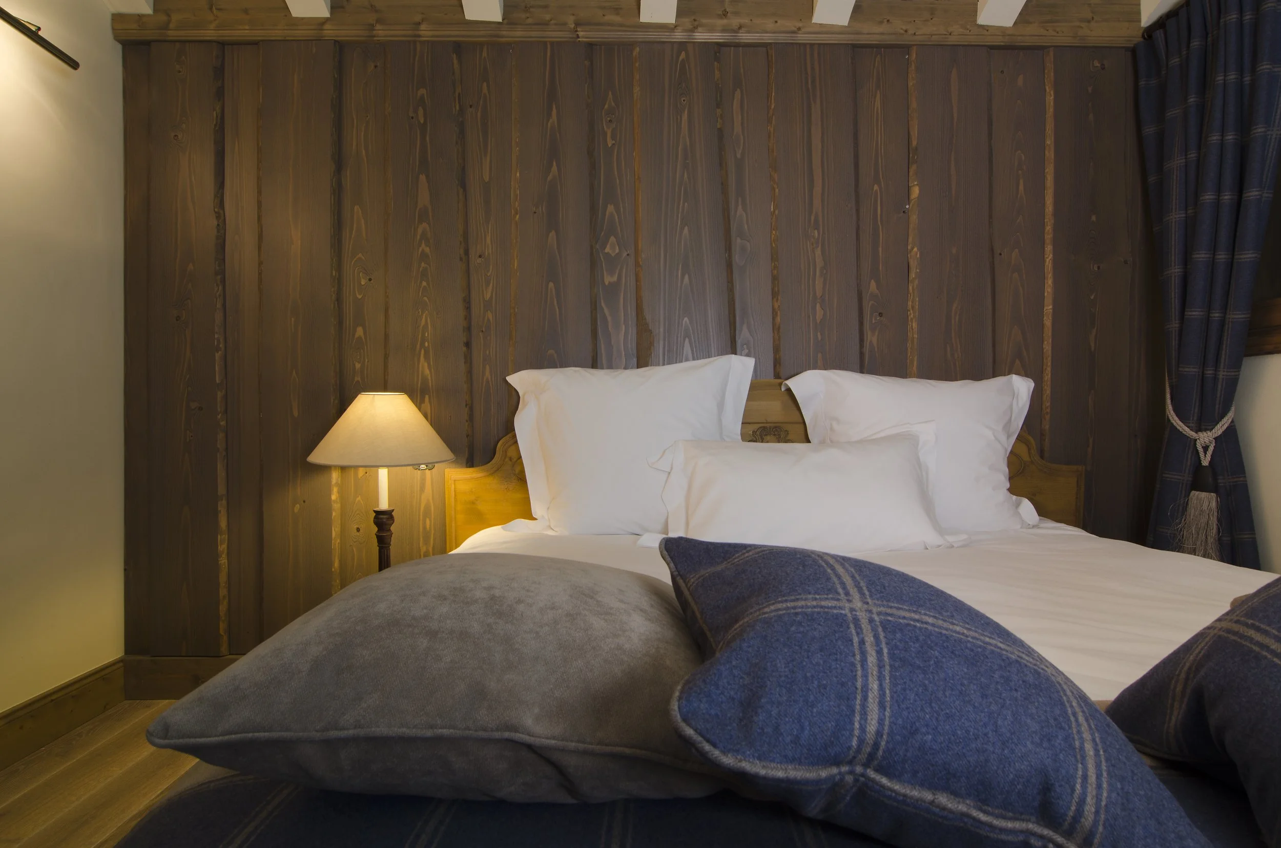 Bett mit weißen Kissen und blauen Kissen vor einer Holzvertäfelung. Seitlich steht eine Lampe, im Hintergrund sind dunkle Vorhänge.
