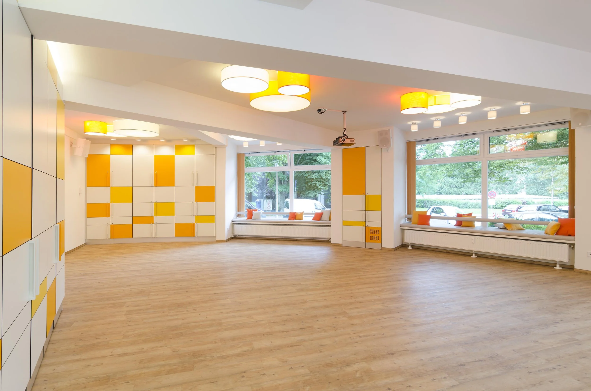 Ein heller, moderner Raum mit großen Fenstern, bunten orange und weißen Wandelementen, Deckenleuchten und einer Holzfußboden. Es gibt Sitzbänke mit bunten Kissen an den Fenstern.