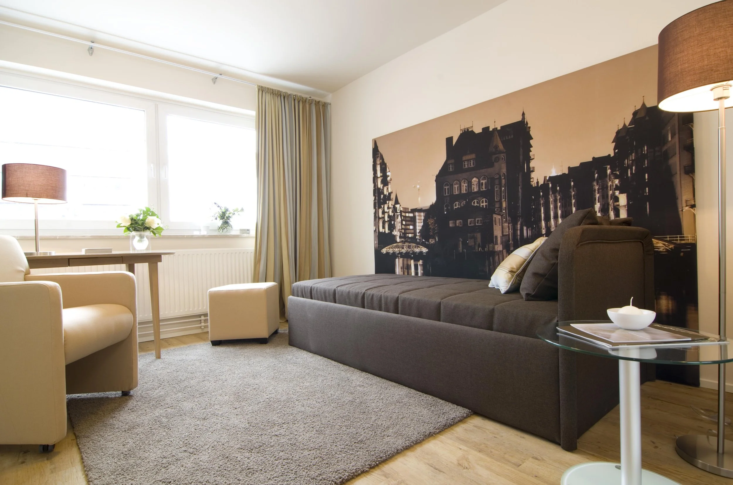 Gemütliches Wohnzimmer mit beige und braun Möbeln, große Fenster mit Vorhängen, Wandbild einer Stadtansicht, Tischlampe, Blumen auf dem Tisch, Teppich auf Holzboden.